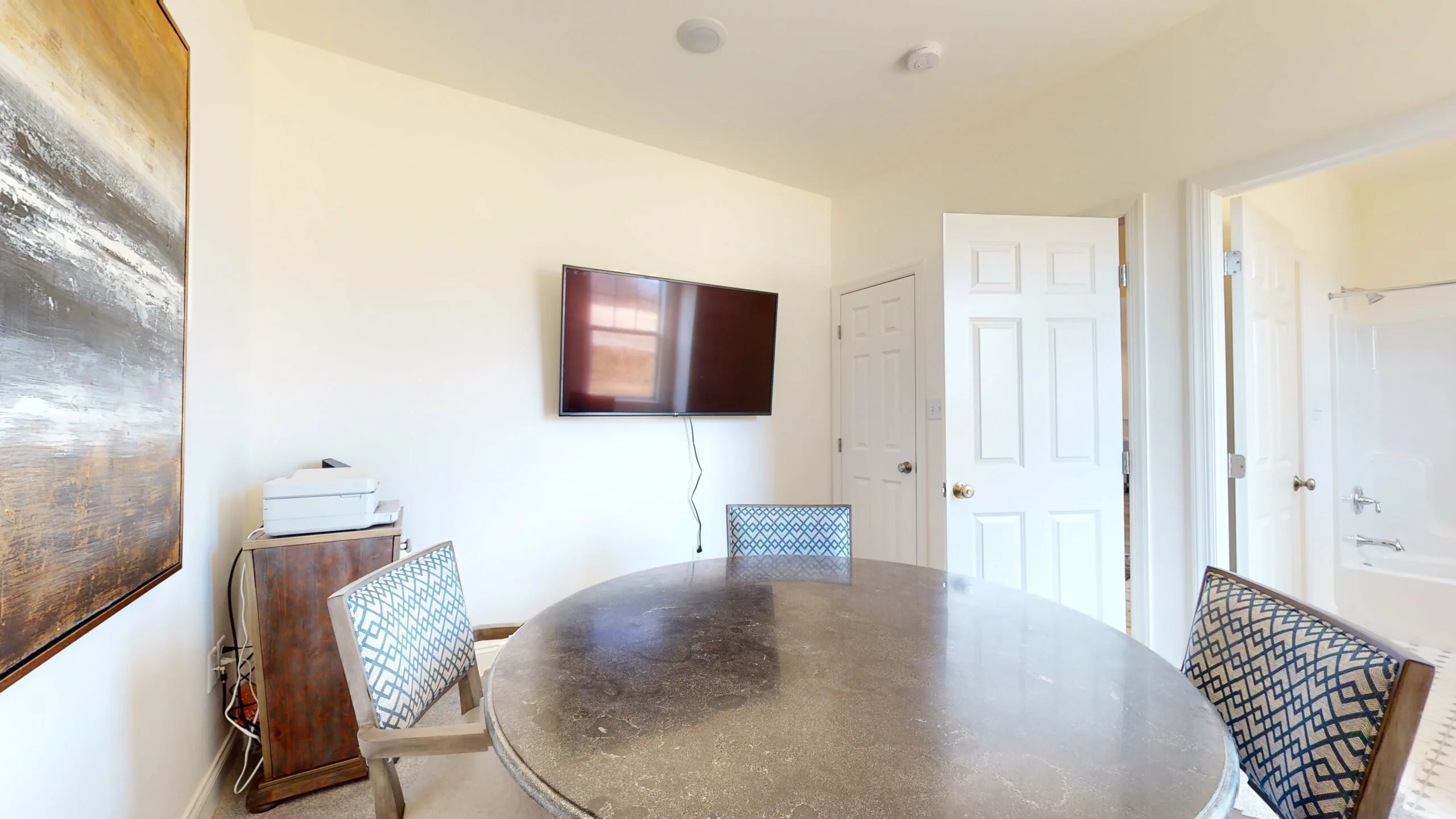 A small, bright room with a round marble table and four patterned chairs. A flat-screen TV is mounted on the wall, and a door is open to a bathroom. A side table holds a printer and stacked papers.