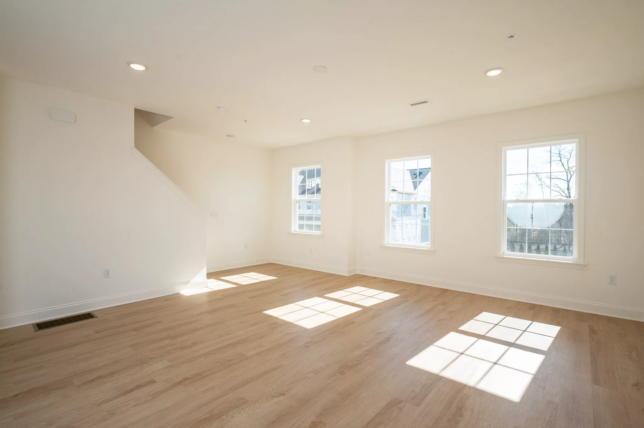 A bright, empty room with light wood floors, white walls, three large windows letting in sunlight, and a staircase in the corner.