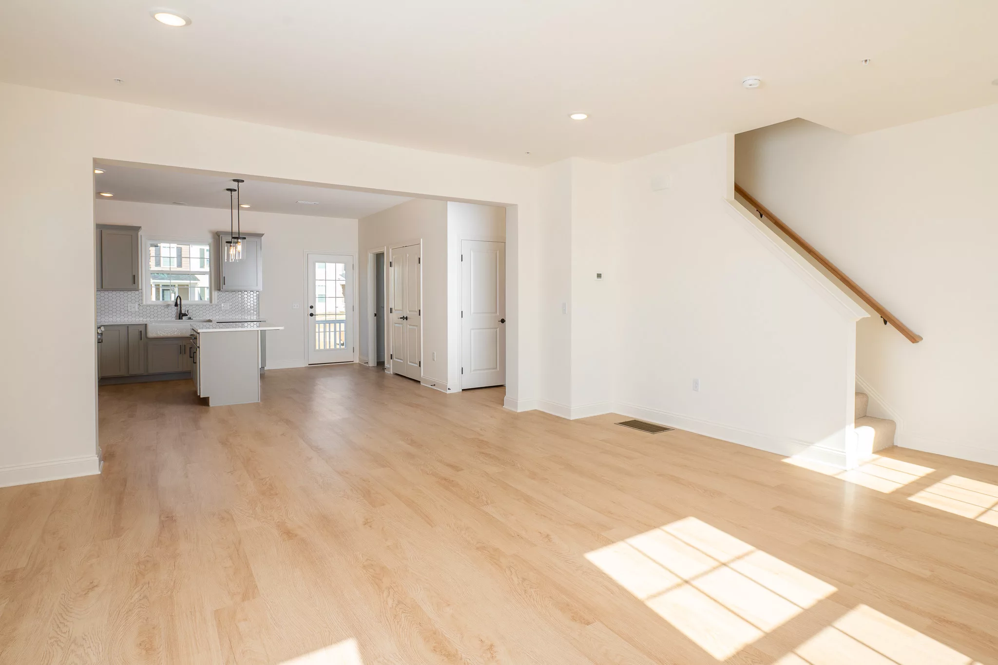 Bright, empty open-plan living area with light wood floors, white walls, and a modern kitchen in the background. A staircase is on the right, and sunlight streams in through large windows.