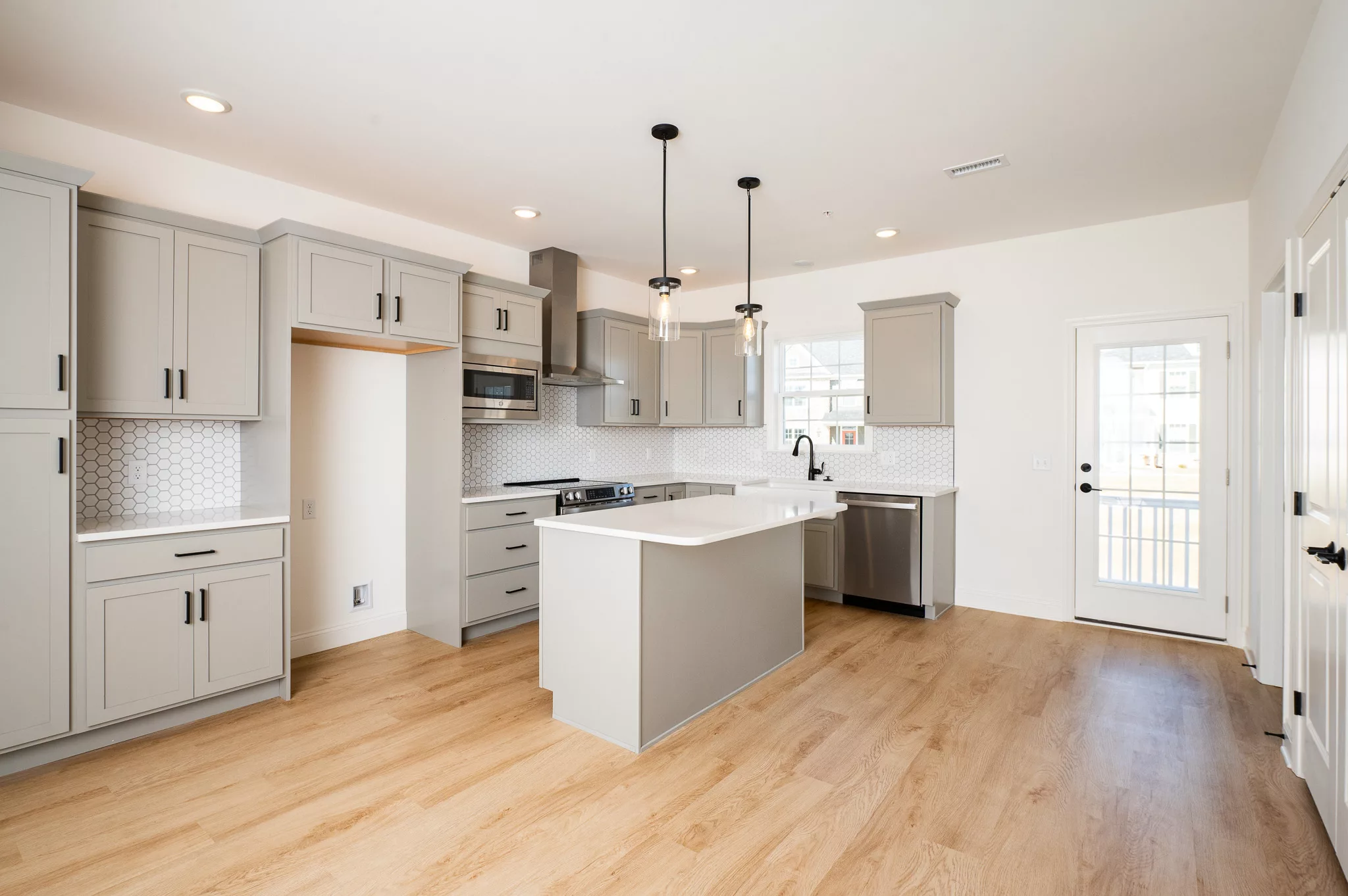 Modern, spacious kitchen with light gray cabinets, white countertops, a center island, stainless steel appliances, pendant lights, white hexagon tile backsplash, and light wood flooring. Natural light enters through a glass door.