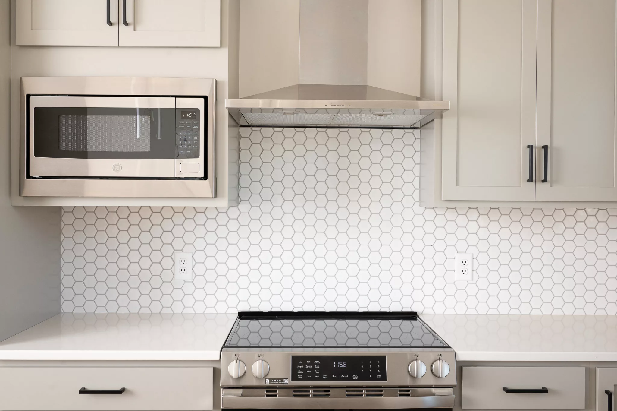 A modern kitchen features a stainless steel microwave, a range hood, and an electric stove with digital controls. The backsplash is white hexagonal tile, and the countertops and cabinets are light-colored.