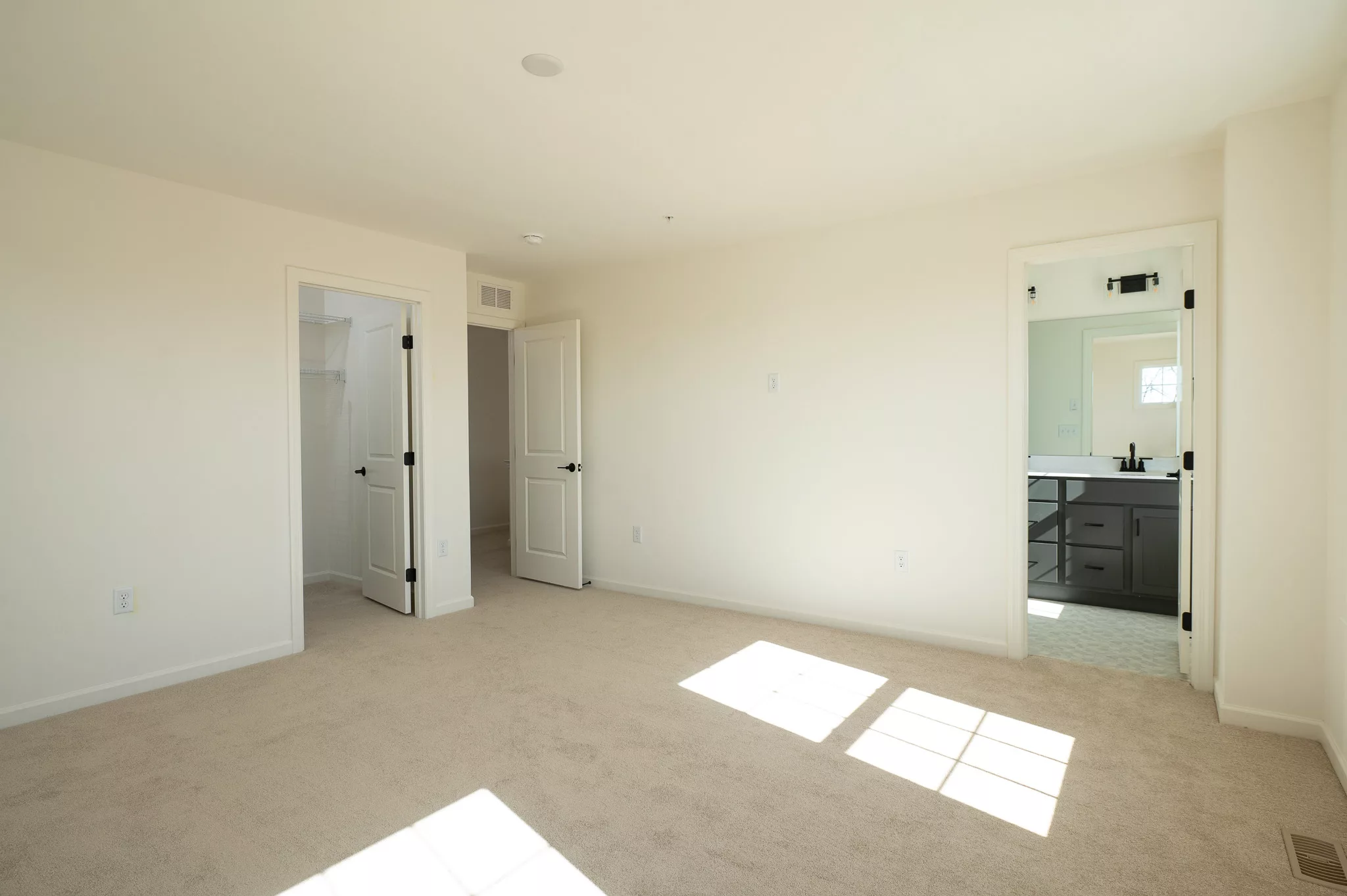 A bright, empty bedroom with beige carpet, cream walls, and three doors: one to a walk-in closet, one to a bathroom with a dark vanity, and one to a hall. Sunlight streams through the window, casting patterns on the floor.