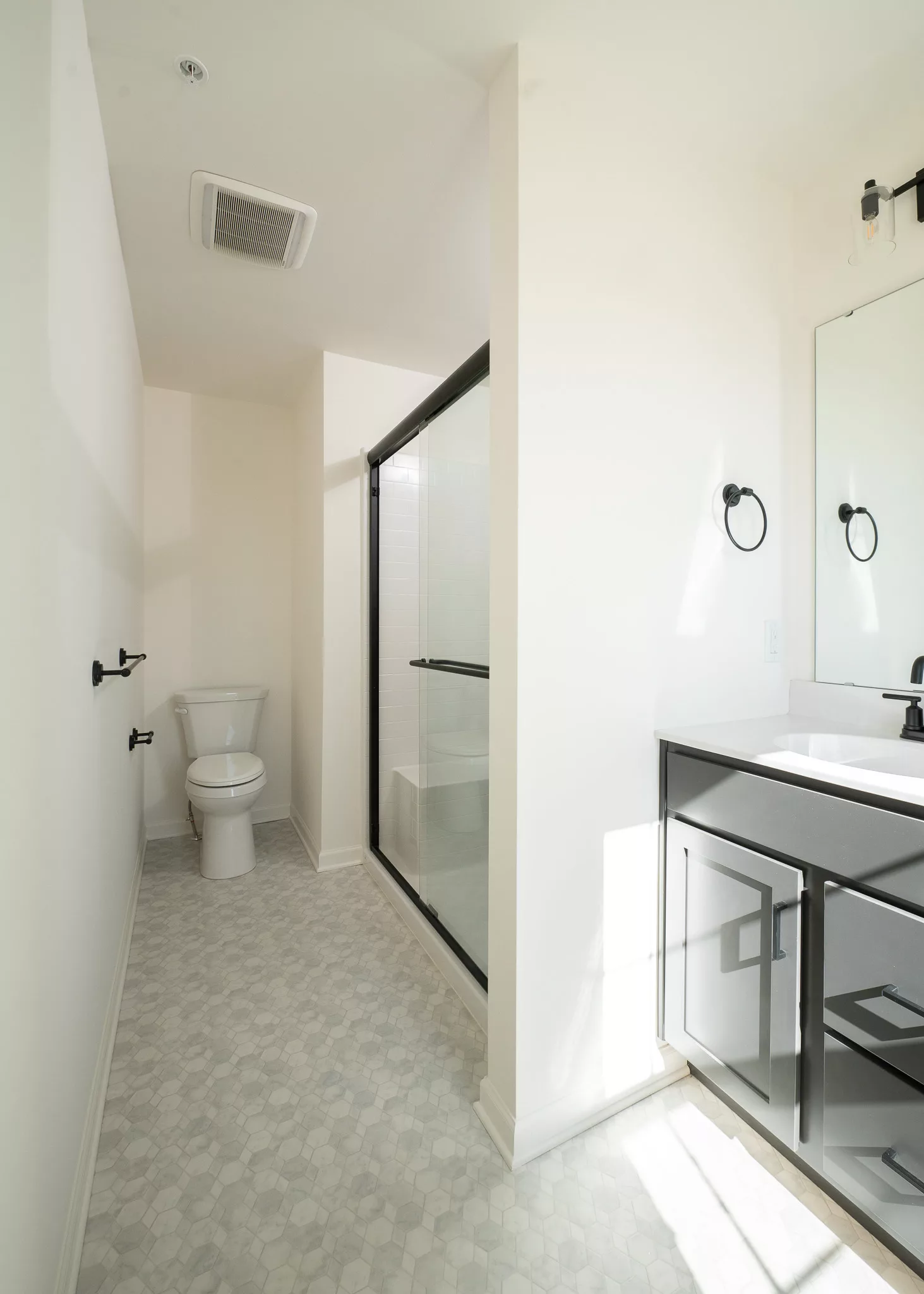 A modern, bright bathroom with white walls, a glass-enclosed shower, a toilet, hexagonal tile flooring, a large mirror, and a vanity with black hardware and sink. Natural light illuminates the space.