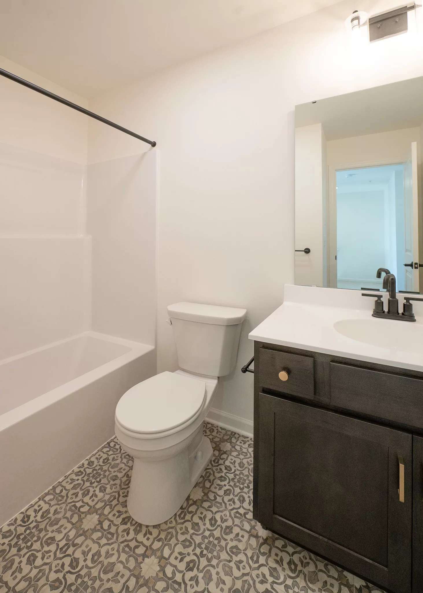 A clean, modern bathroom with a white bathtub and shower, a toilet, a dark wood vanity with a sink and mirror, and patterned tile flooring. The walls are white and lighting is bright.