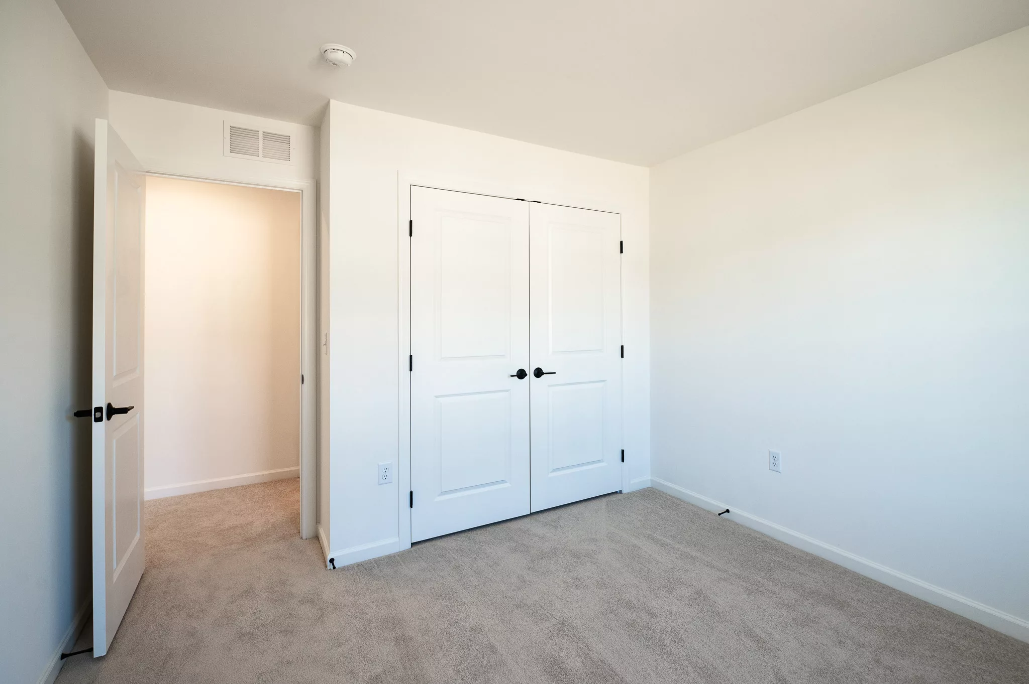 A small, empty room with beige carpet, white walls, an open white door, and a closed double closet with white doors and black handles. The room is well-lit with natural light.