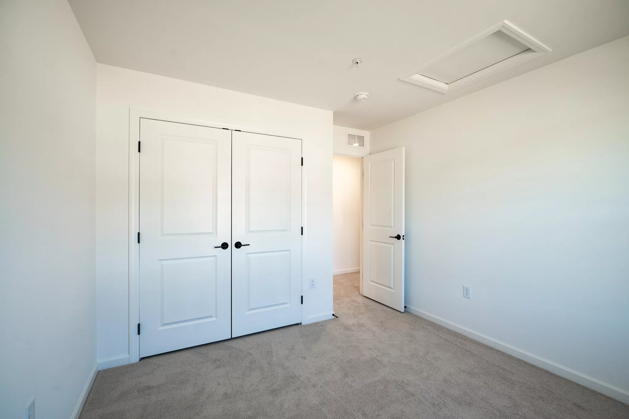 A small, empty room with beige carpet, white walls, a double-door closet, a single door leading to a hallway, and a ceiling attic access panel. The space is well-lit with natural light.