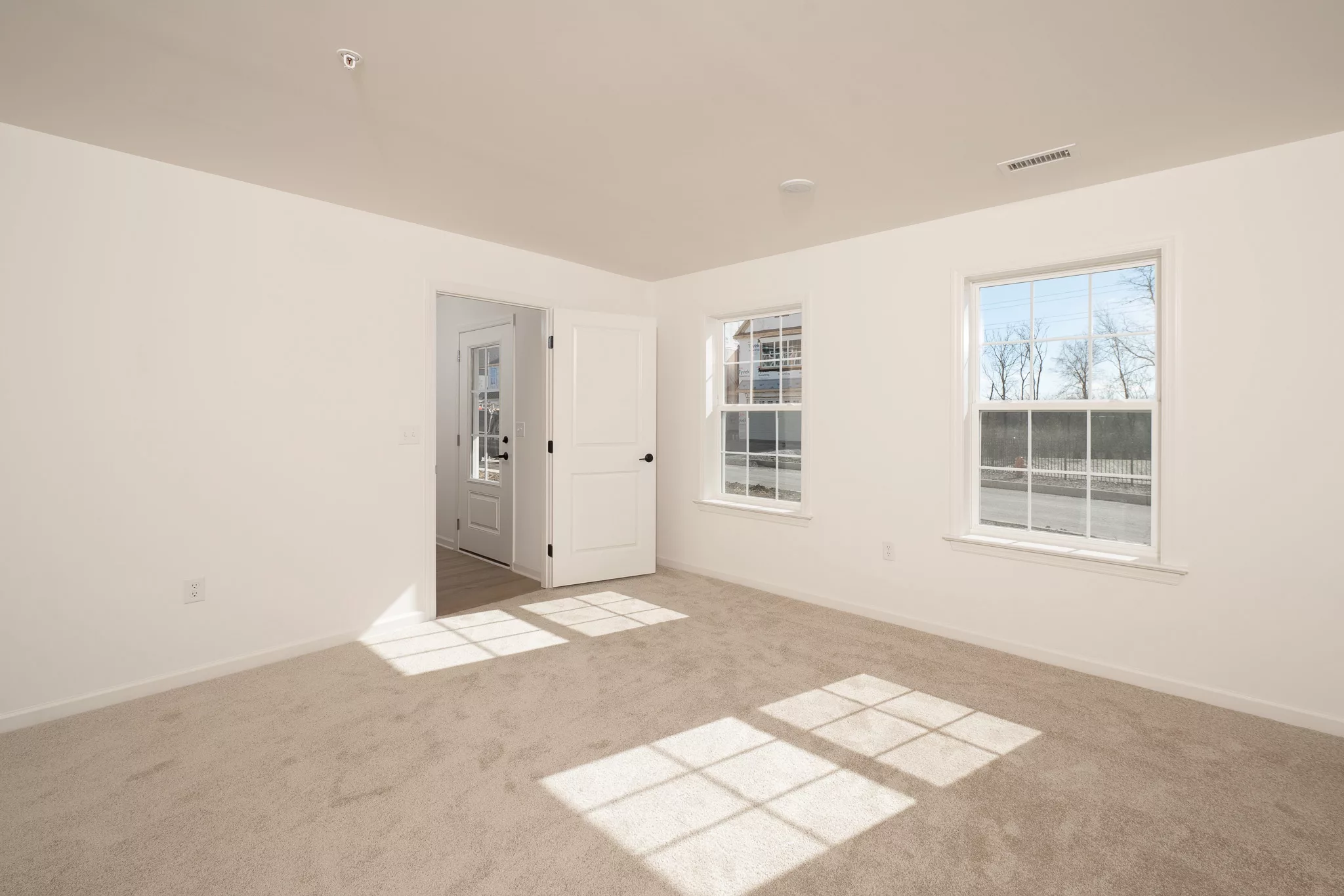 A bright, empty room with beige carpet, white walls, two large windows letting in sunlight, and a white door leading to another room. Shadows from the windows fall onto the carpet.