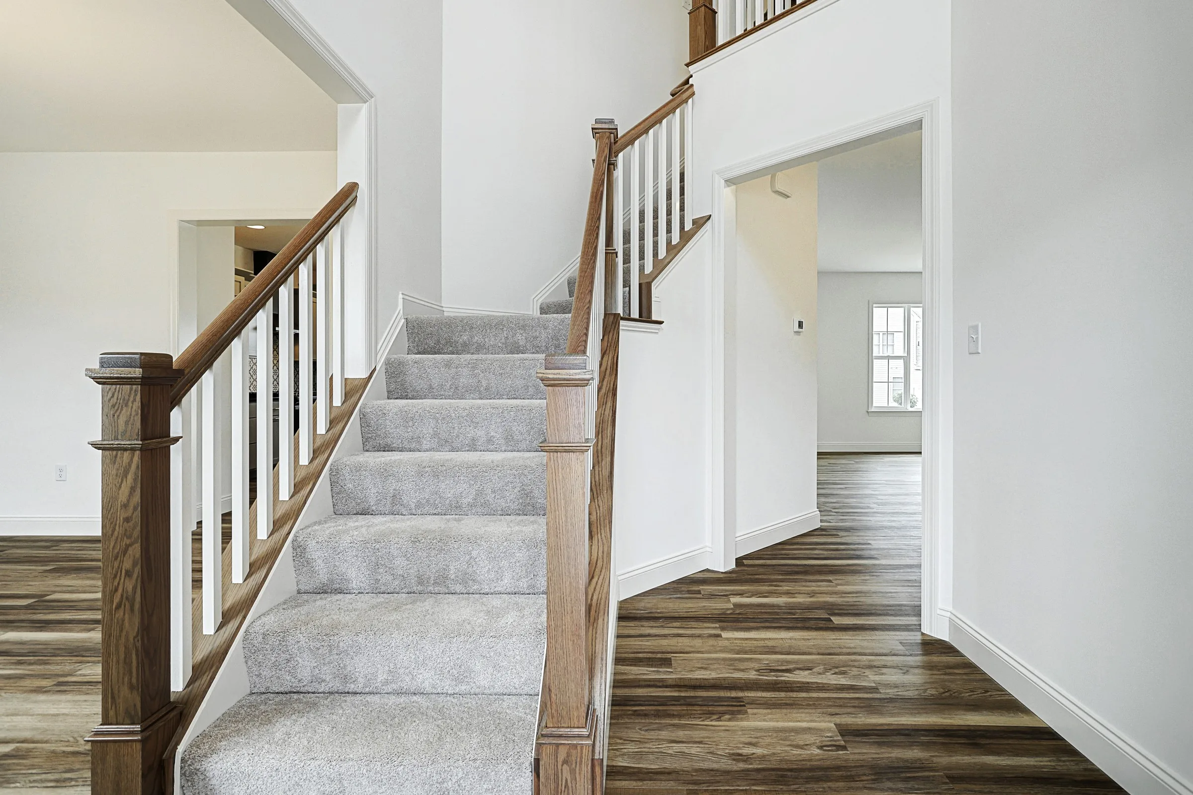 A modern home interior featuring a staircase with wooden handrails and carpeted steps, white walls, and wood-style flooring. The space is bright with natural light coming through large windows.