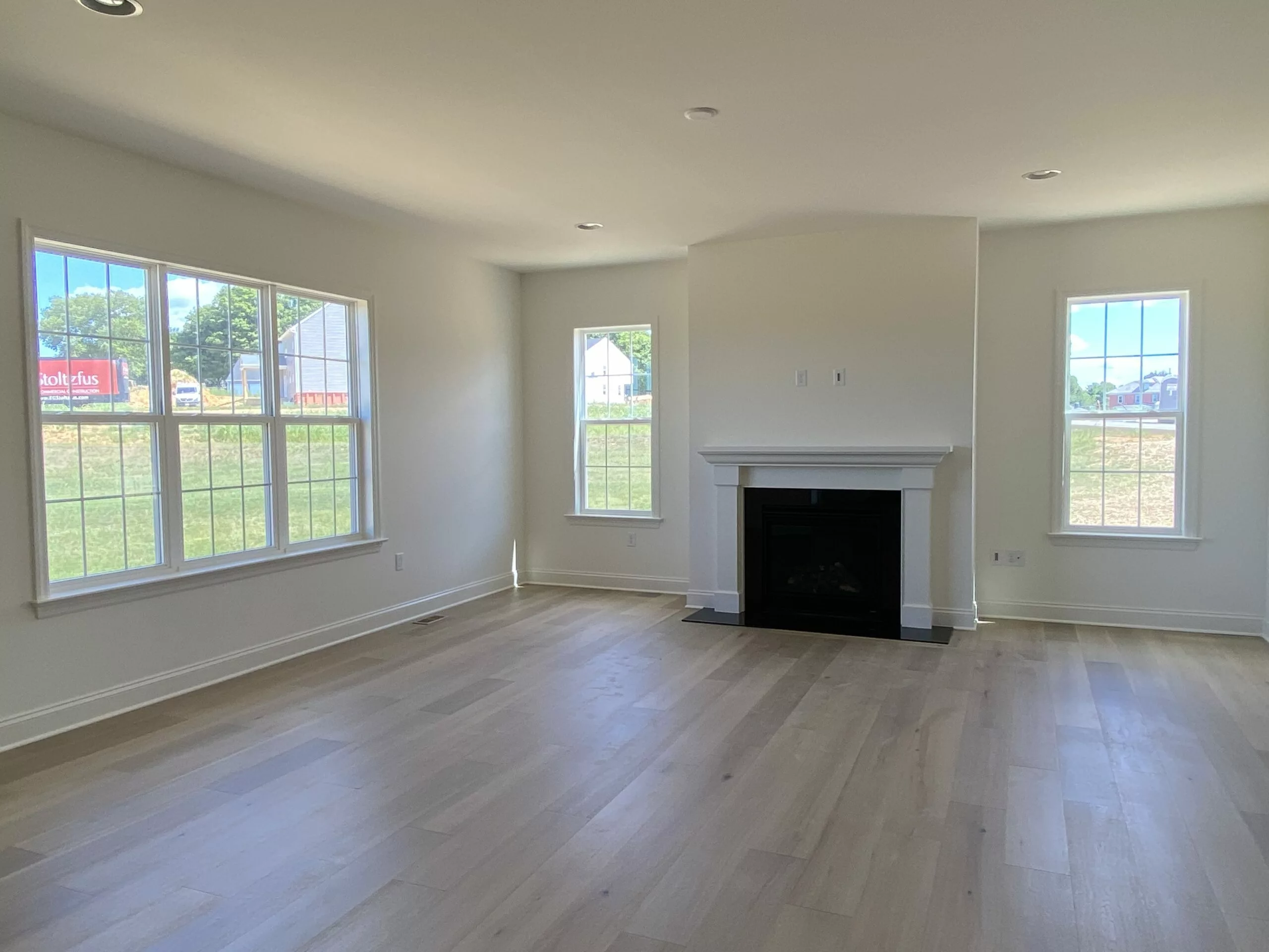 Bright, empty living room with light wood floors, large windows letting in natural light, and a white fireplace centered on the far wall. Green grass and buildings are visible outside.