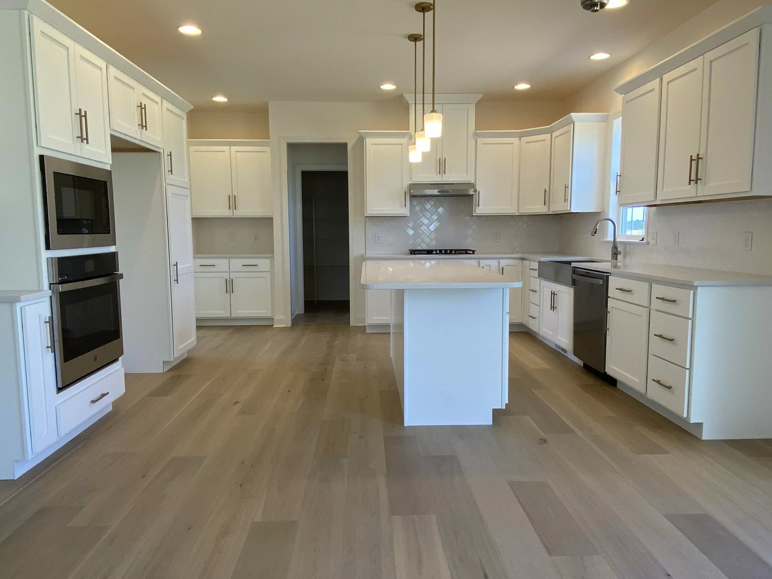 Modern kitchen with white cabinets, stainless steel appliances, a center island with pendant lights, light wood flooring, and neutral walls. The room is spacious and well-lit with recessed lighting.