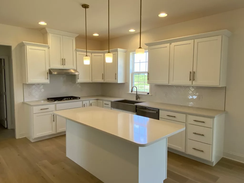 Modern kitchen with white cabinets, stainless steel appliances, an island with a quartz countertop, pendant lights, light wood flooring, and a window above the sink letting in natural light.