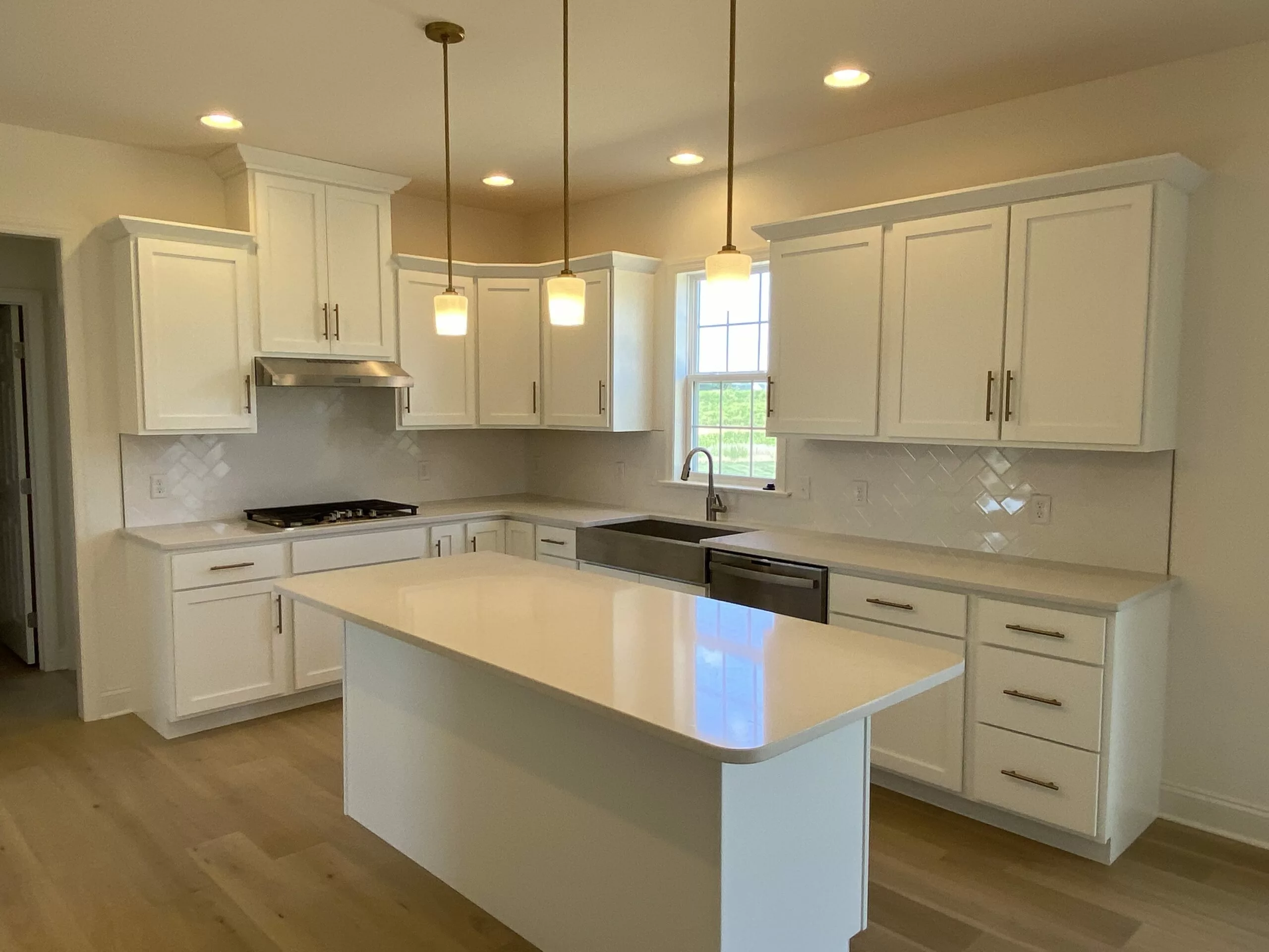 Modern kitchen with white cabinets, stainless steel appliances, an island with a quartz countertop, pendant lights, light wood flooring, and a window above the sink letting in natural light.