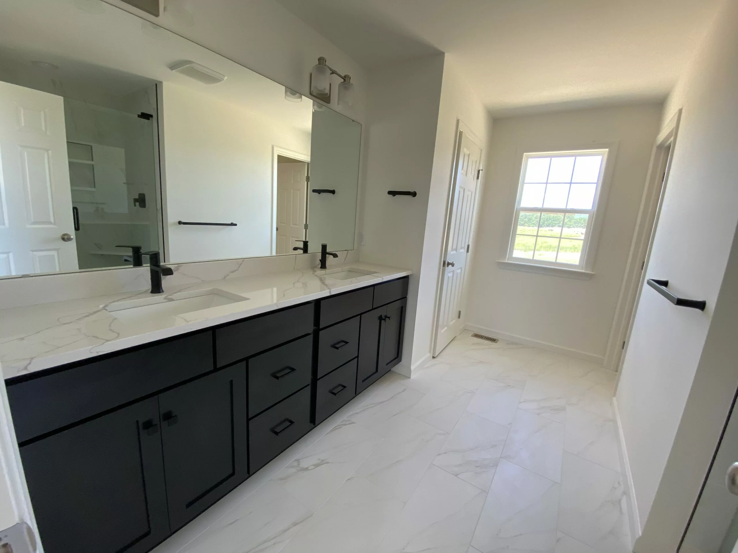 Modern bathroom with white marble tile flooring, a large mirror above a double-sink vanity with black cabinets and fixtures, and a window letting in natural light.