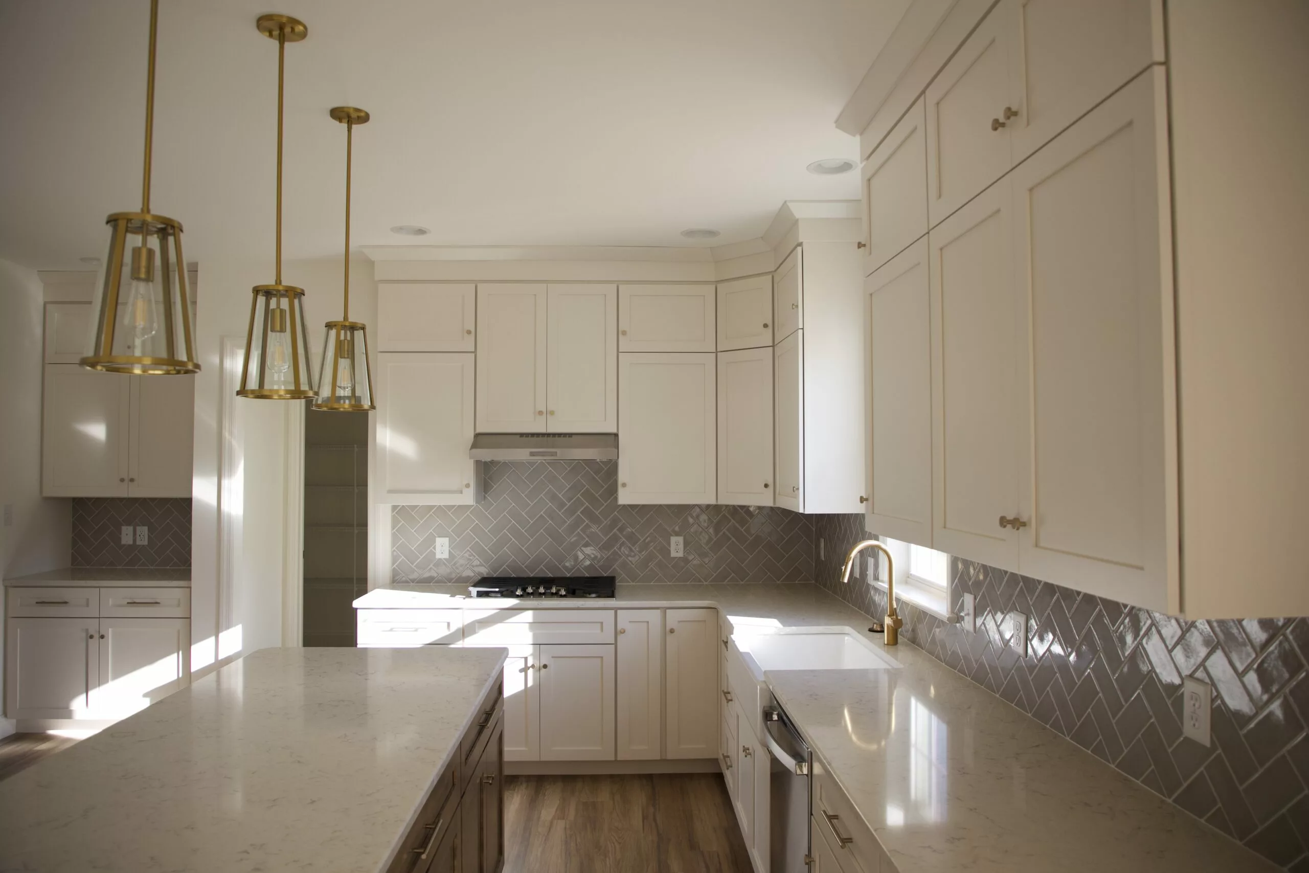 Bright, modern kitchen with white cabinets, marble countertops, gold pendant lights, stainless steel appliances, and a herringbone-patterned tile backsplash. Sunlight streams in, highlighting the clean and elegant design.