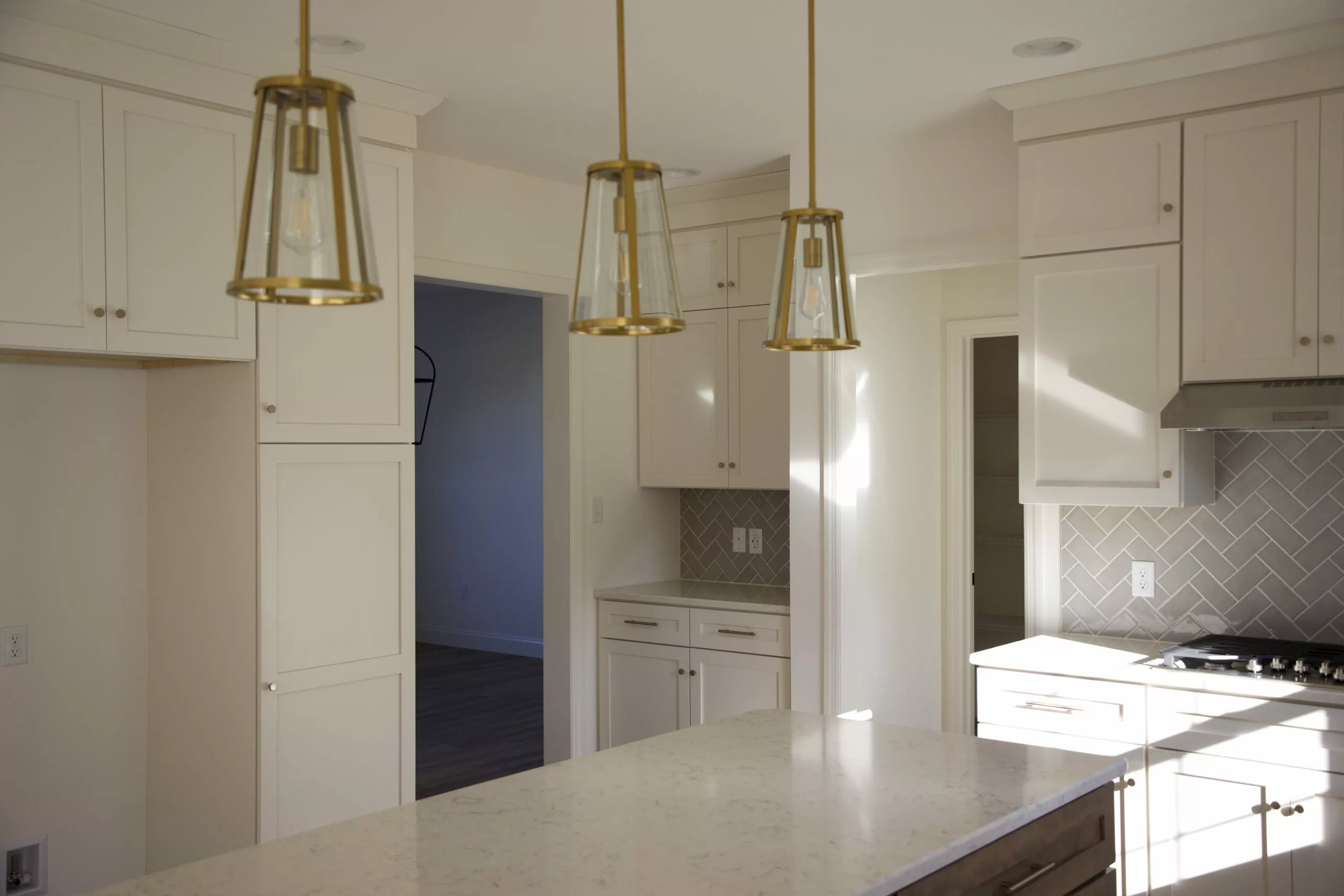 Bright modern kitchen with white cabinets, a marble island, gold pendant lights, beige herringbone backsplash, and sunlight streaming through the windows, creating a warm and inviting atmosphere.