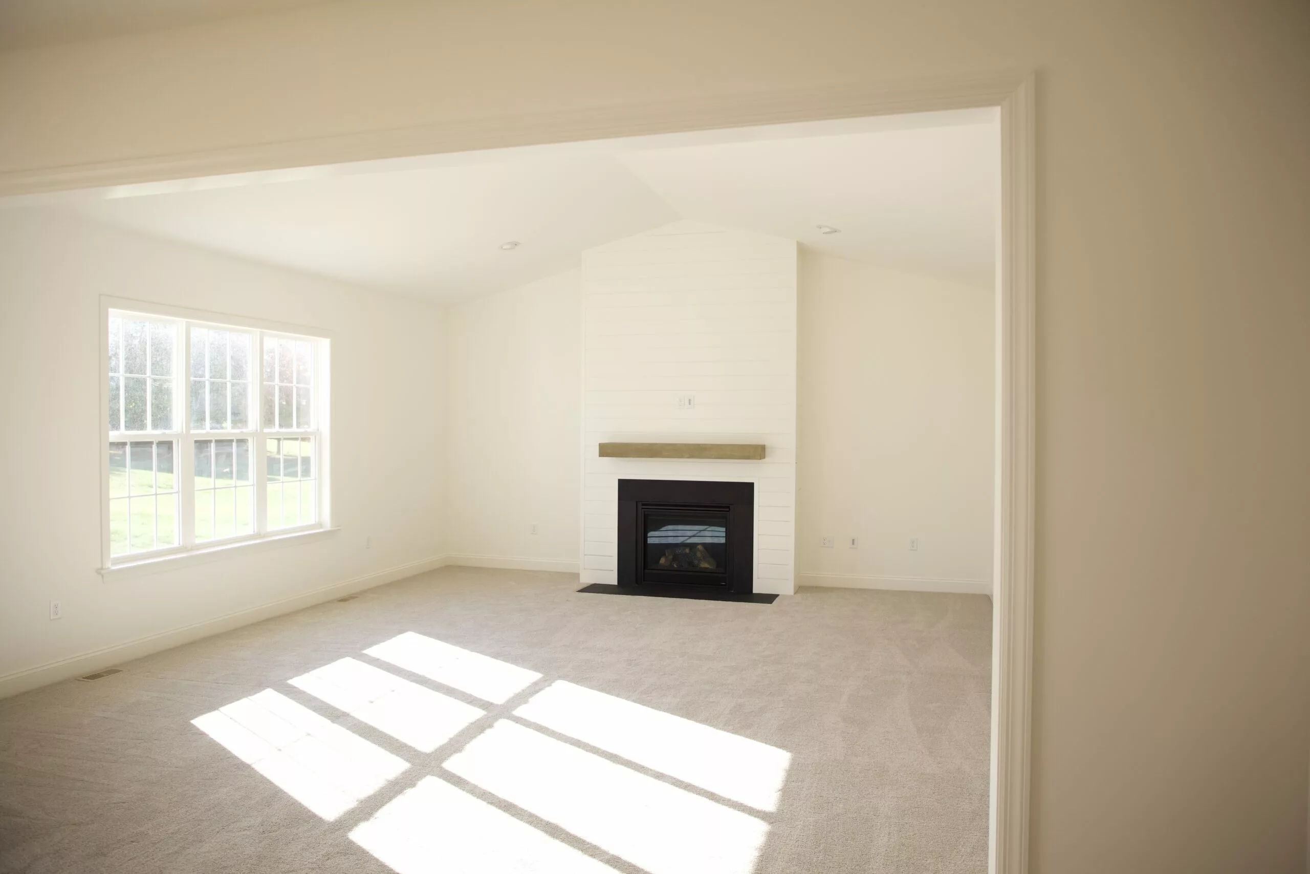 A bright, empty living room with cream-colored walls, carpeted floor, large window letting in sunlight, and a fireplace with a wooden mantel centered on the far wall.