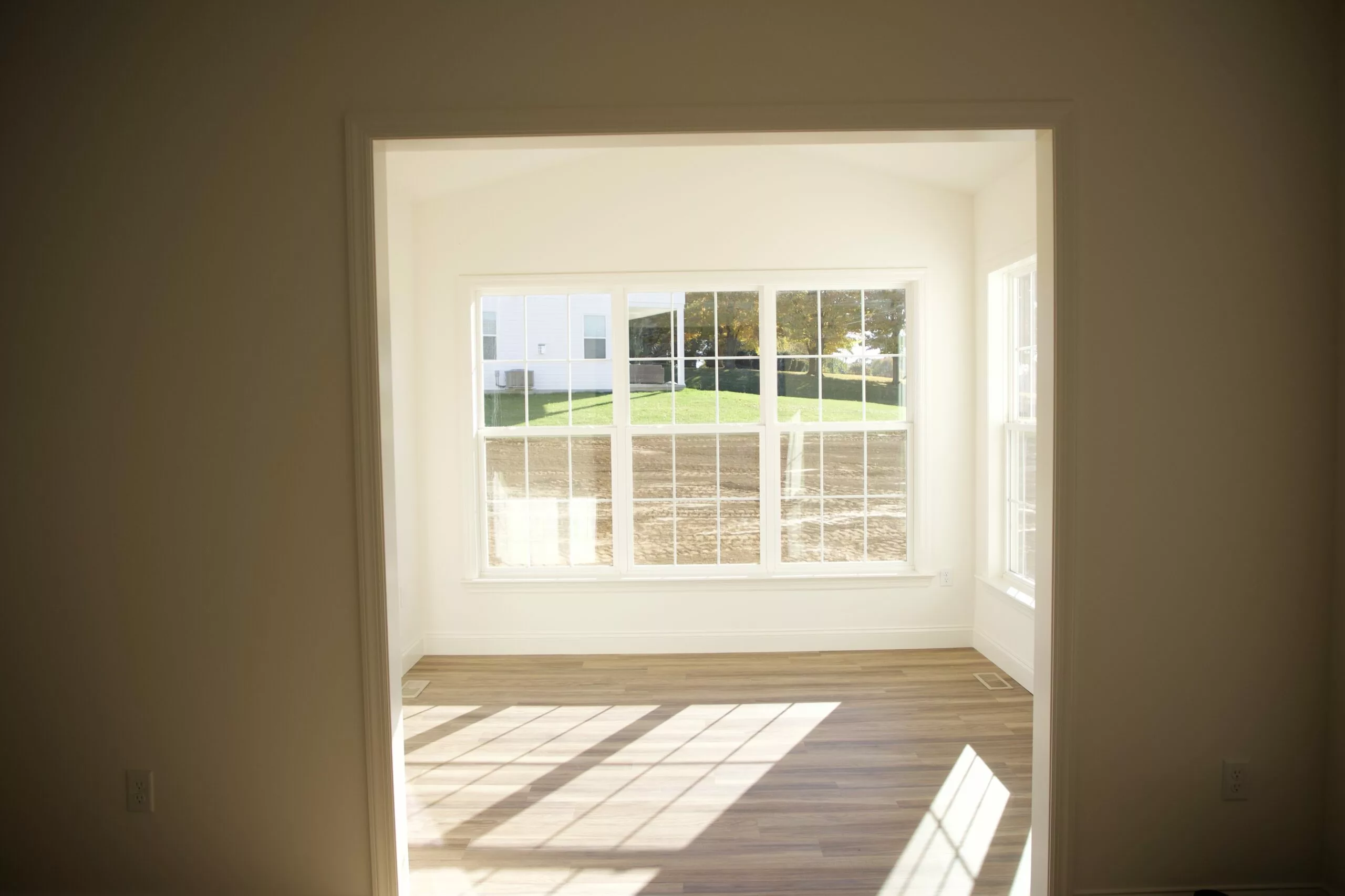 A bright, empty room with light wood flooring and large windows letting in sunlight. The windows offer a view of grass, trees, and a neighboring building outside.