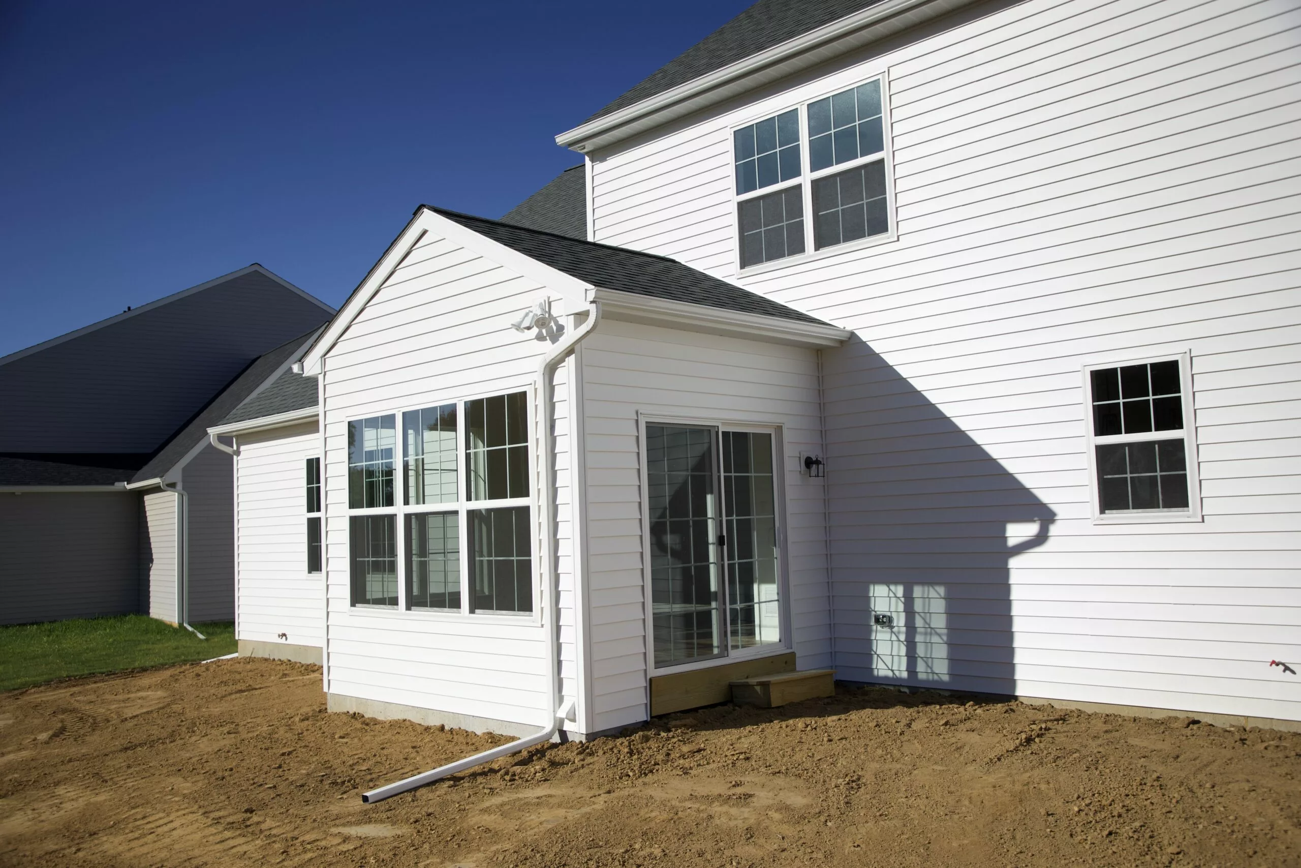 A newly-built white house with large windows and a sliding glass door opens to a yard with bare soil, under a clear blue sky. The house has clean siding and no landscaping yet.