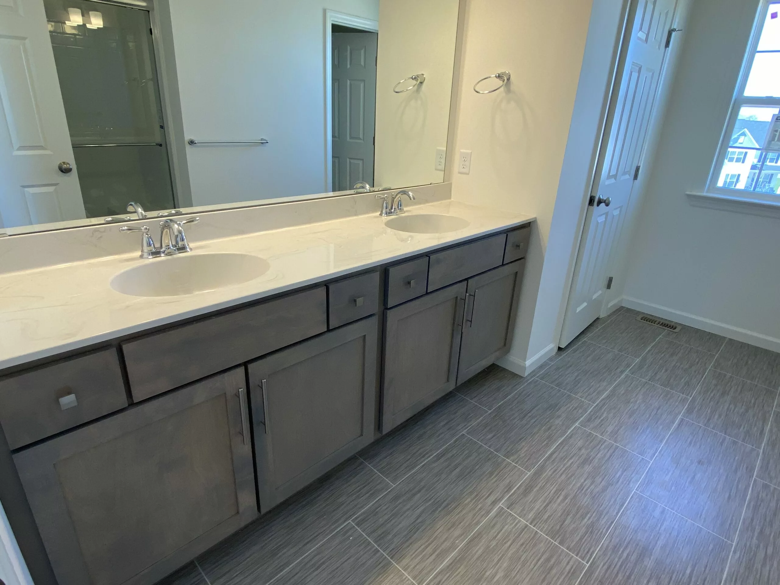 Modern bathroom with a double sink vanity, large mirror, gray tiled floor, and neutral walls. There is a glass shower door to the left and a window to the right letting in natural light.