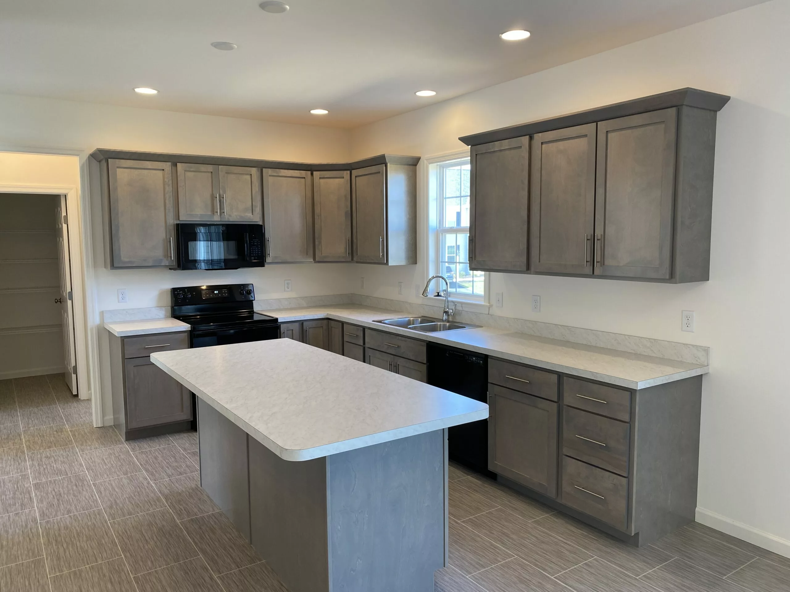 Modern kitchen with gray cabinets, white countertops, a center island, black appliances, a window above the sink, tile flooring, and recessed lighting. A doorway to the left leads to a walk-in pantry.