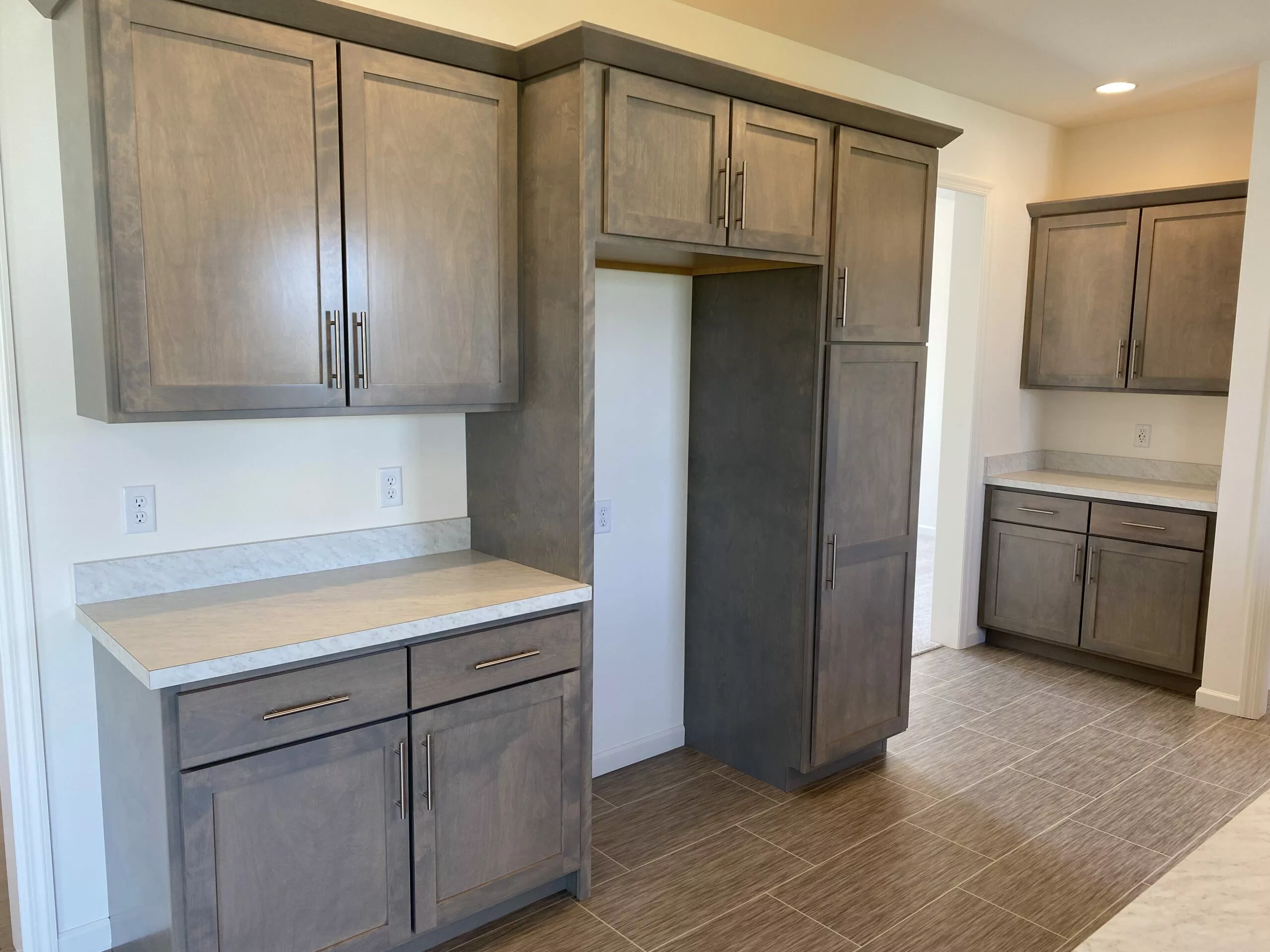 A modern kitchen with light brown wooden cabinets and drawers, beige countertops, and brown tiled flooring. There is an empty space for an appliance between the cabinets. Another counter area is visible in the background.