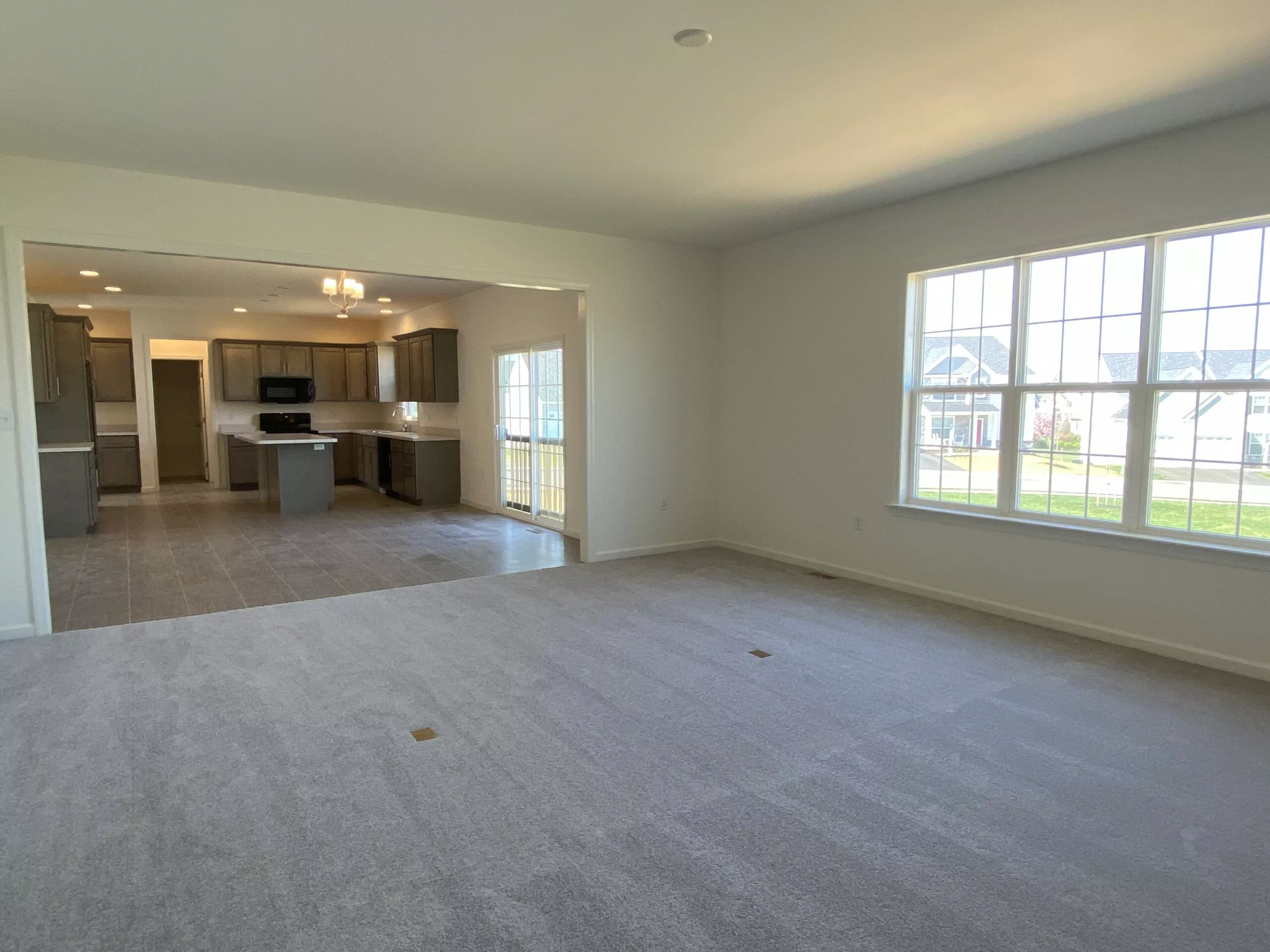 Spacious, unfurnished living room with light gray carpet, large windows letting in natural light, and an open view into a modern kitchen with tiled floor, cabinets, and stainless steel appliances.