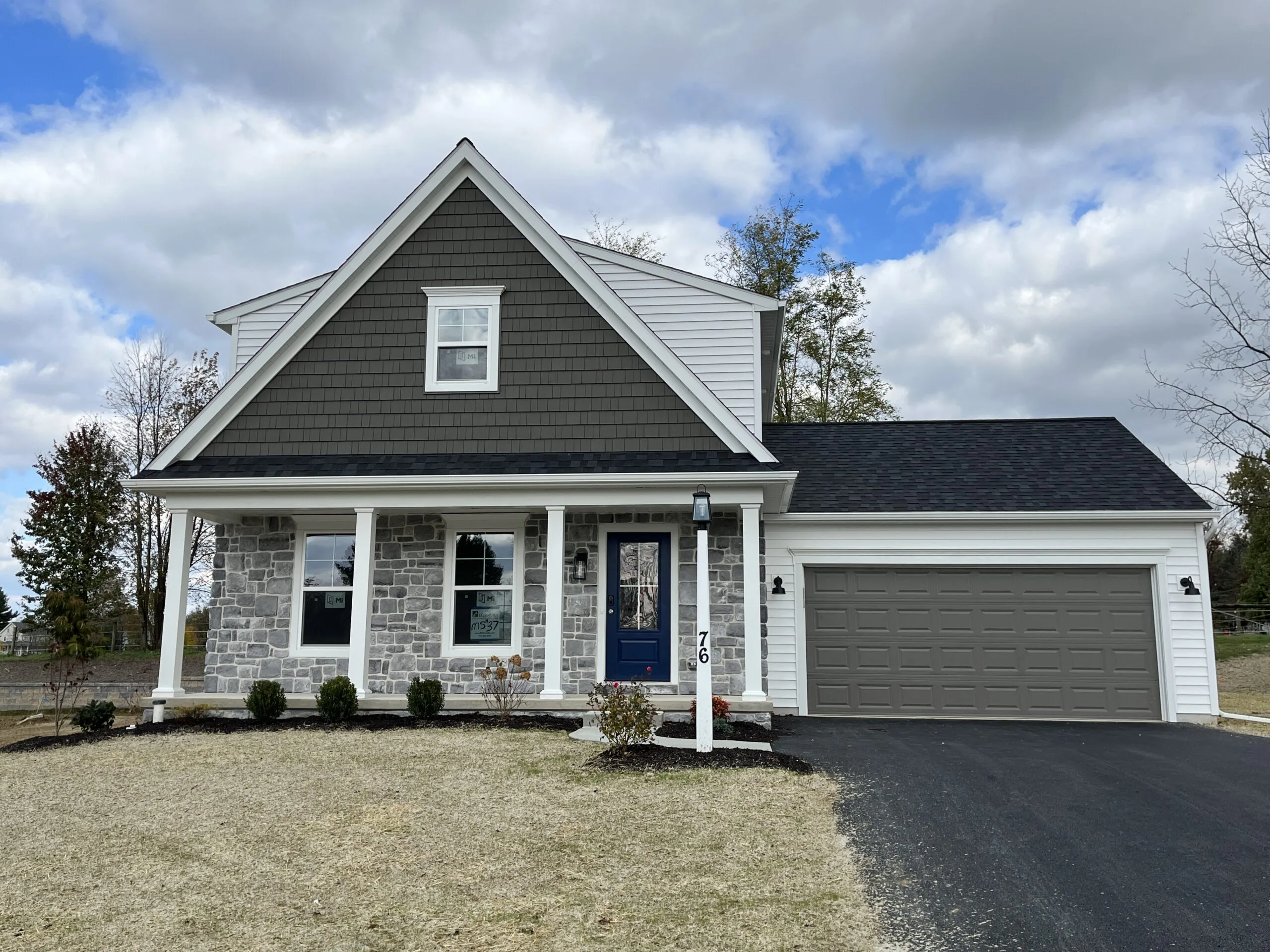 A modern suburban house with gray siding and stone facade, a front porch with white pillars, blue front door, and attached two-car garage. The yard has minimal landscaping and a paved driveway. Cloudy sky in the background.
