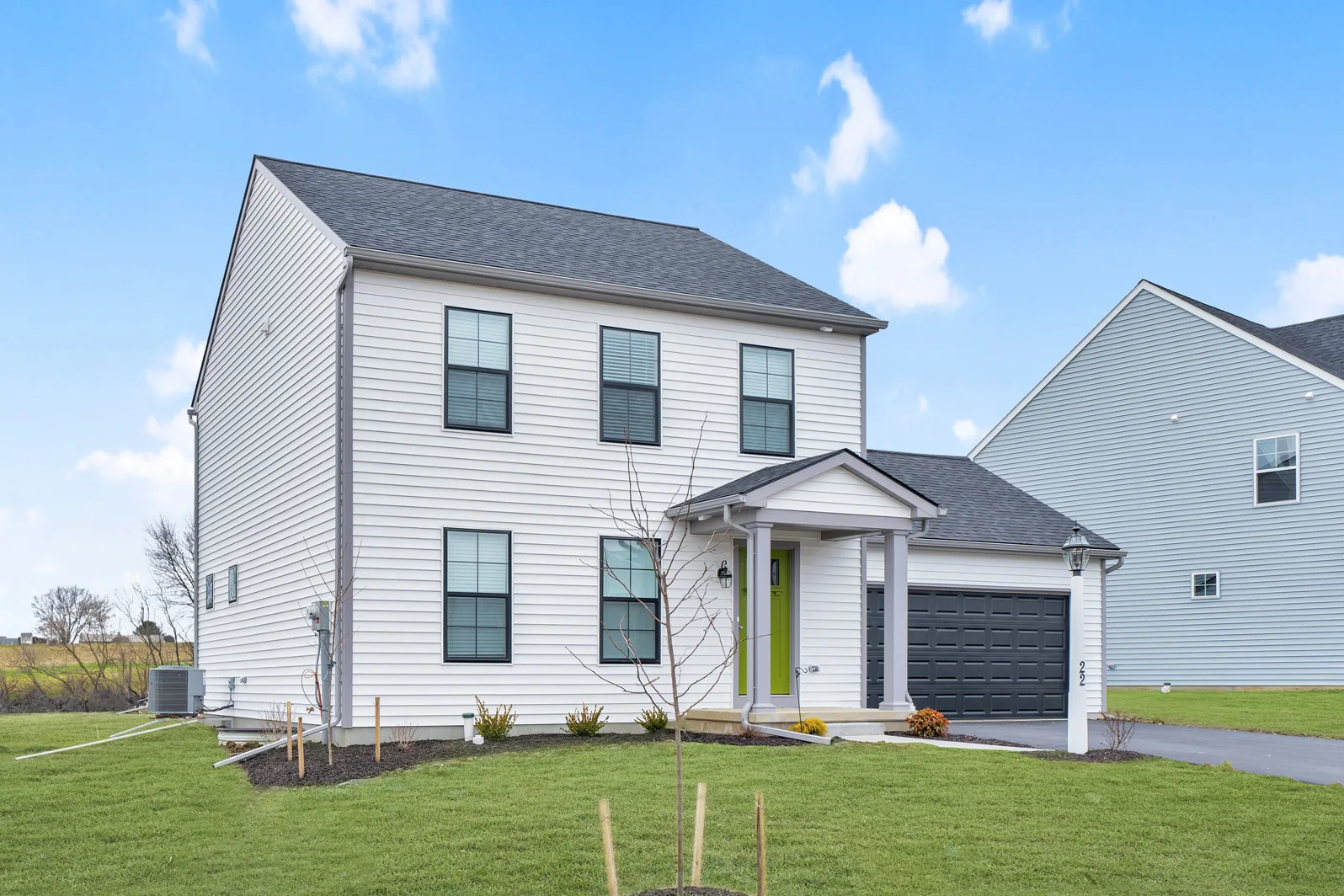 Two-story modern house with white siding, black-framed windows, a bright green front door, attached two-car garage, and a small covered porch. The house sits on a lawn with young trees and clear blue sky overhead.