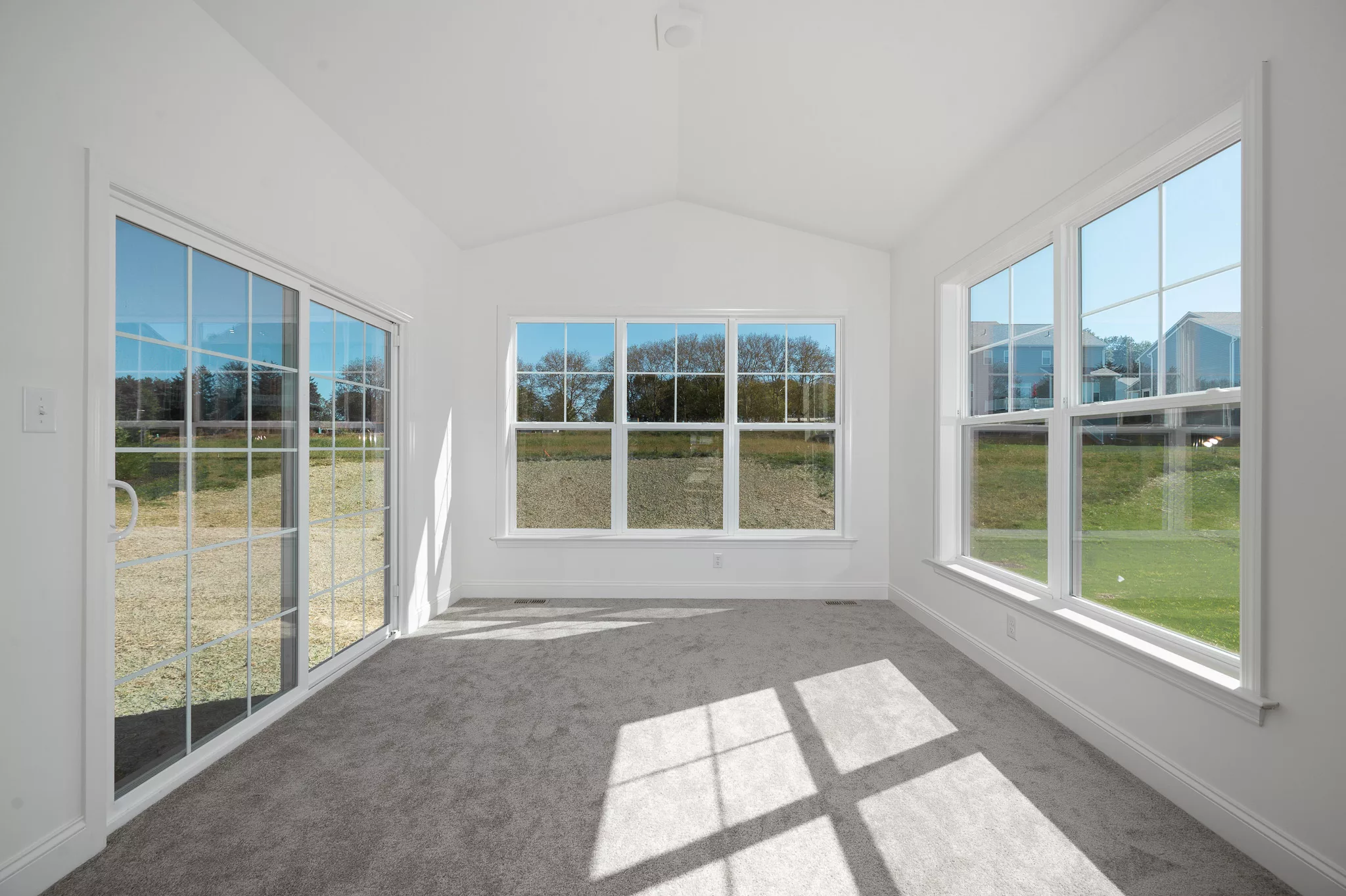 A bright, empty sunroom with large windows on three sides, light gray carpet, and white walls. Sunlight streams in, casting sharp shadows on the floor. A glass sliding door opens to a grassy outdoor area.