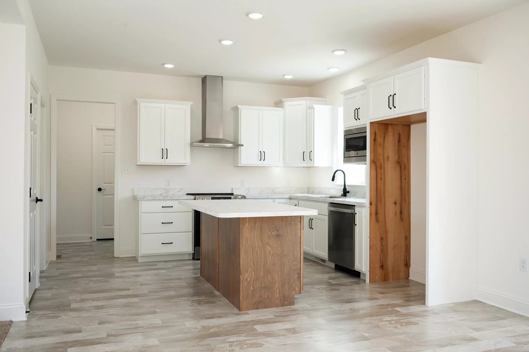Modern kitchen with white cabinets, stainless steel appliances, a marble countertop, a wooden island, light wood flooring, and recessed lighting. There is an empty space for a refrigerator on the right.