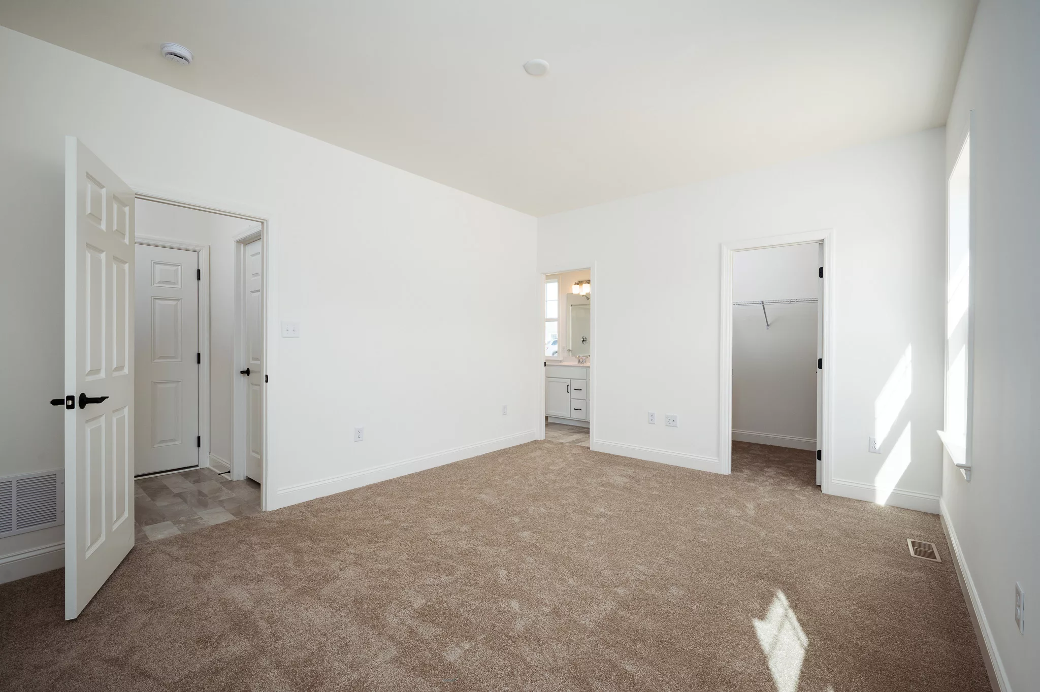 A bright, empty bedroom with beige carpet, white walls, open doors leading to a bathroom and a walk-in closet, and natural light coming through windows on the right.