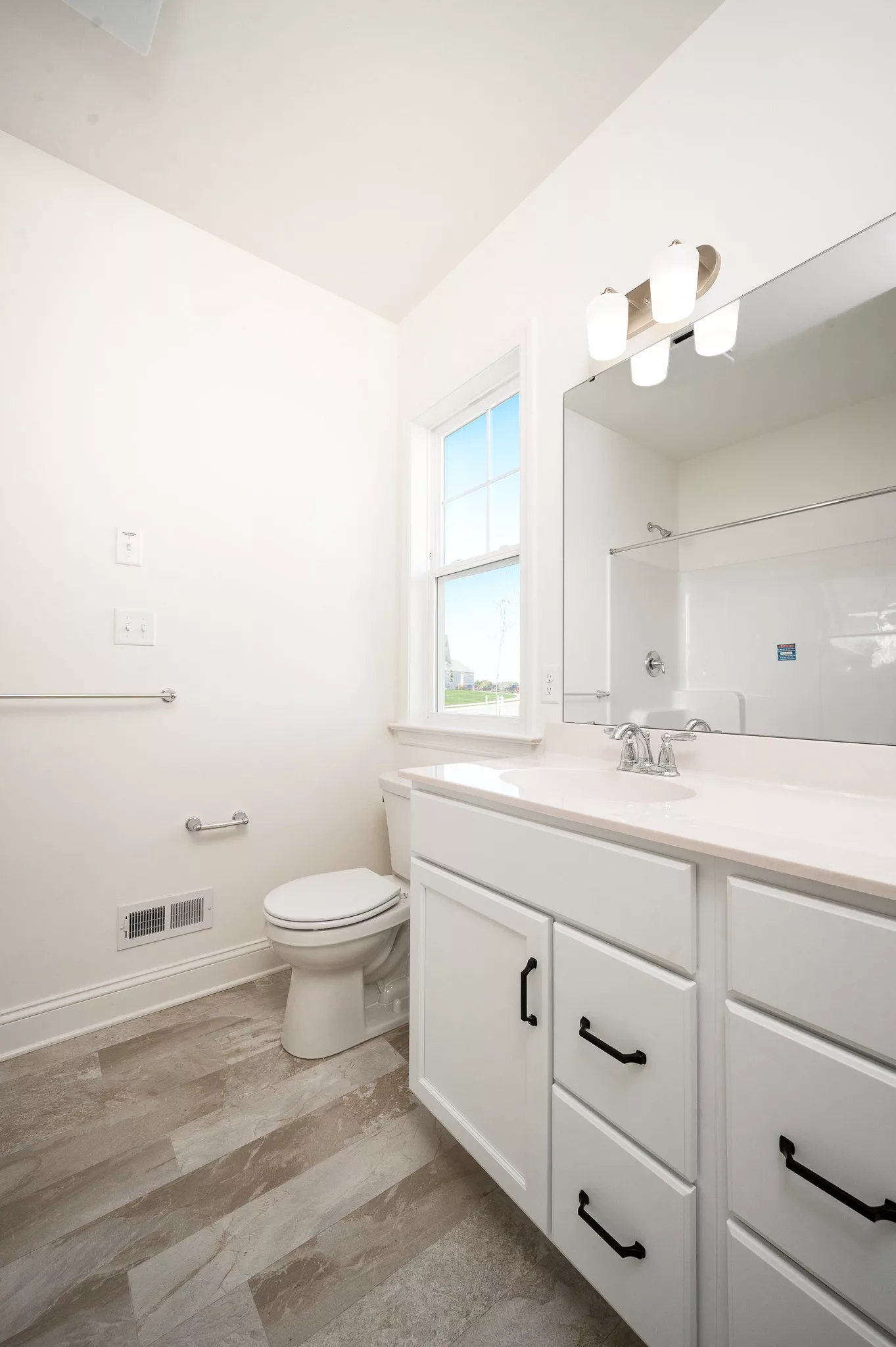 Bright, modern bathroom with white walls, a large mirror above a white vanity with black handles, a toilet next to a window, and a walk-in shower visible in the background.