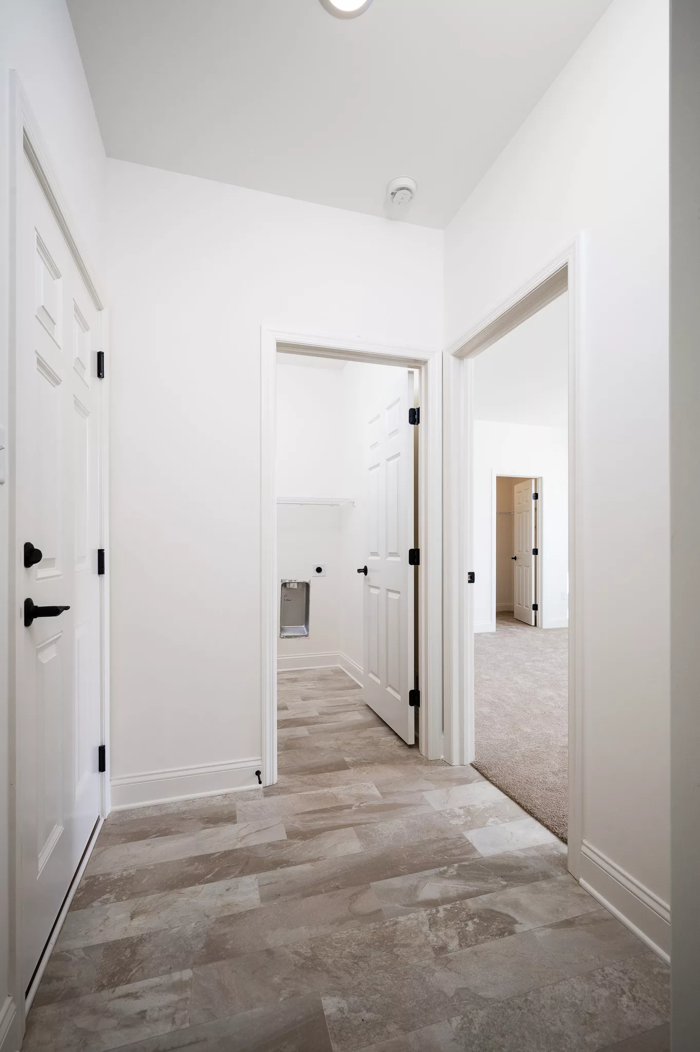 A hallway with light-colored tile flooring and white walls, featuring three open doors leading to different rooms. One room has carpet flooring, and another has shelving and a towel rack.