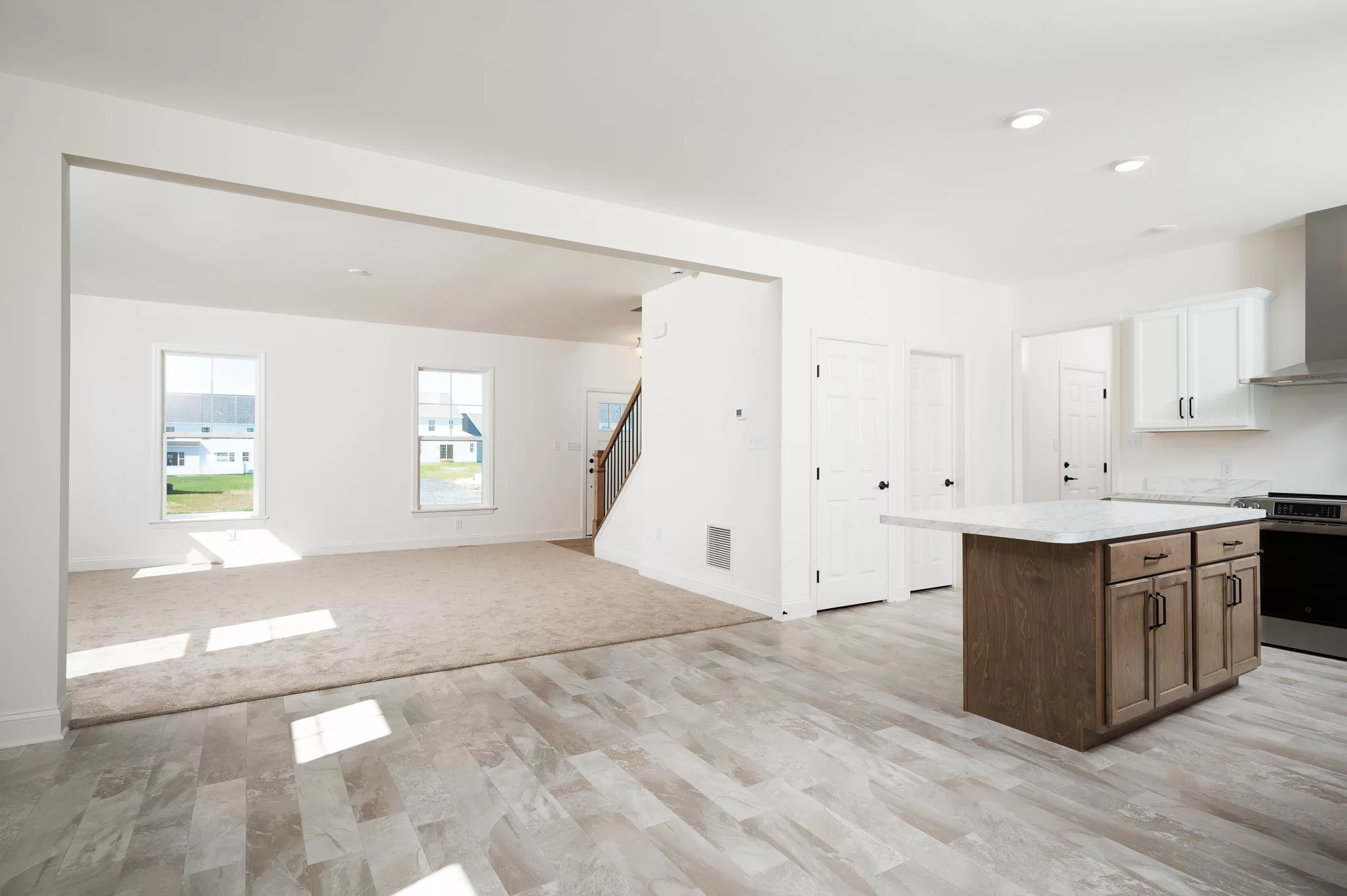 Bright, open-concept living space with large windows, beige carpet in the living area, and tile flooring in the kitchen. Features a kitchen island, white walls, and a staircase near the entryway.