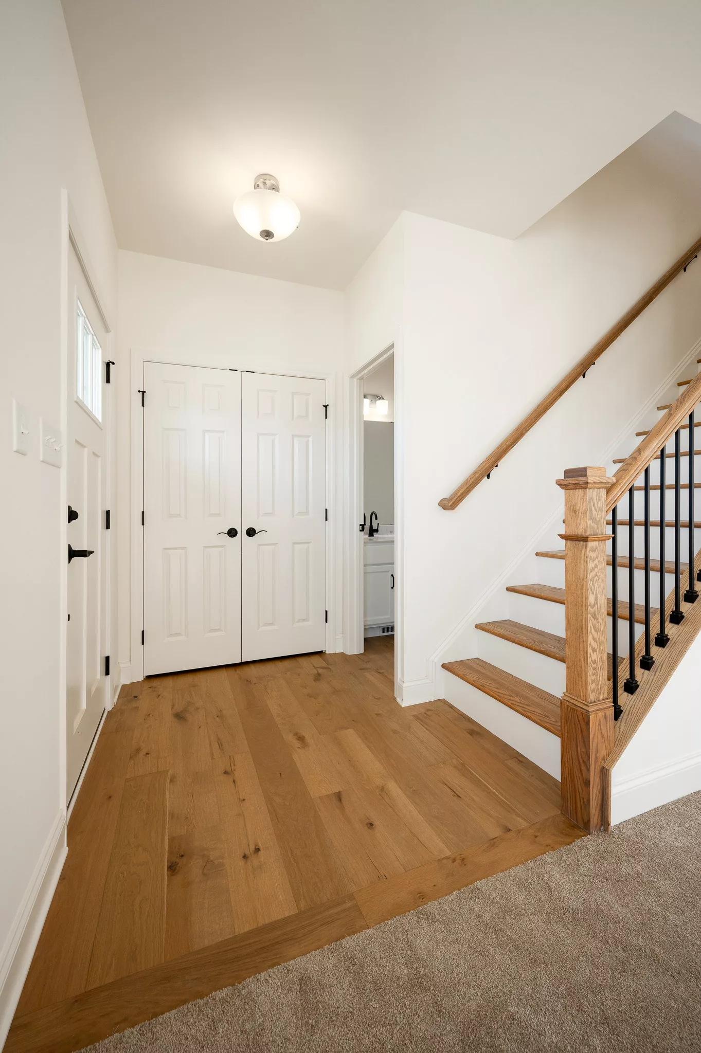 A bright, modern entryway with light wood floors, white walls, a staircase with wooden handrail and black balusters, double closet doors, and a simple ceiling light fixture.