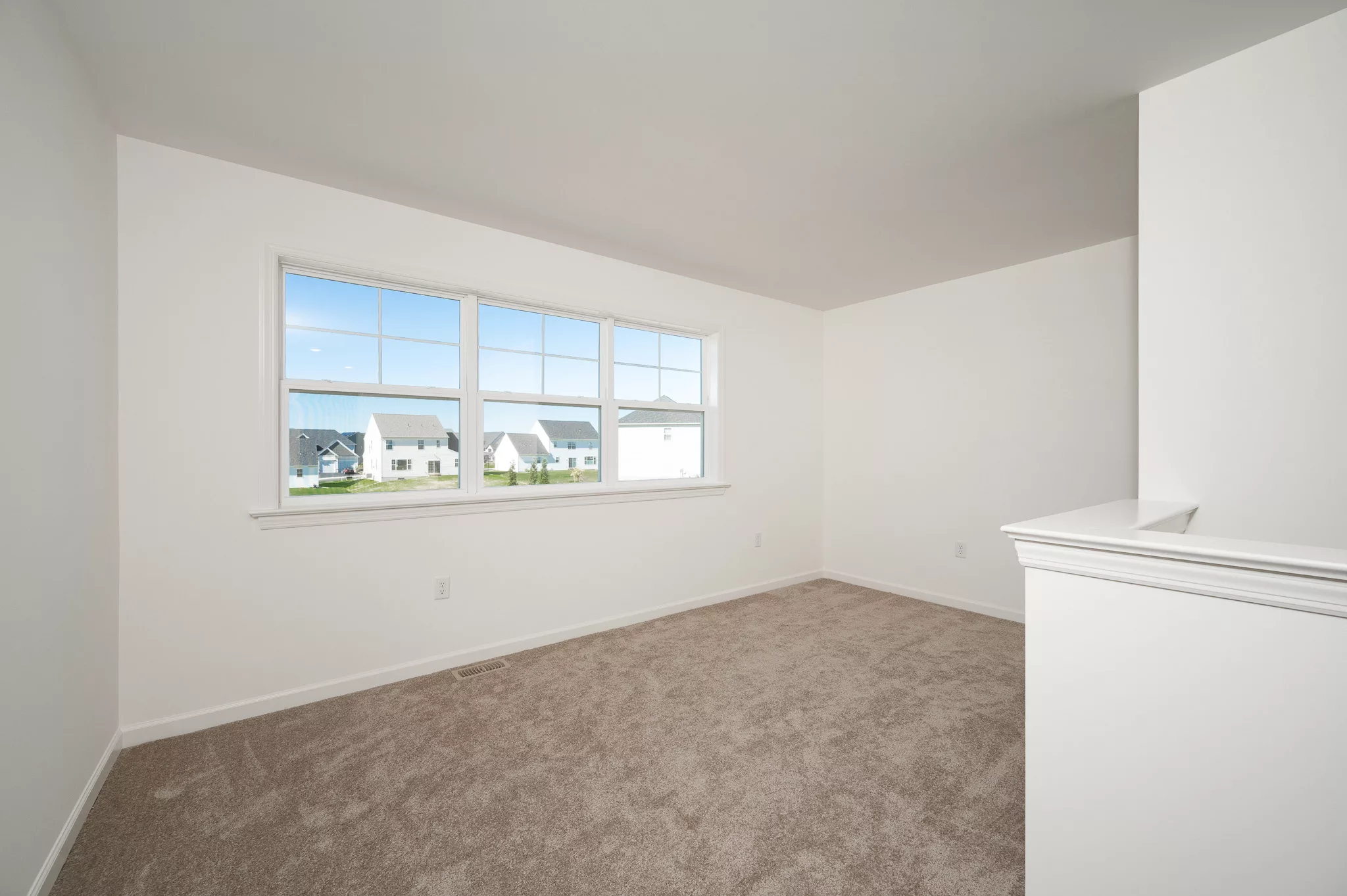 Bright, empty room with beige carpet, white walls, and a large window overlooking suburban houses on a sunny day. The room appears freshly painted and clean, with natural light filling the space.