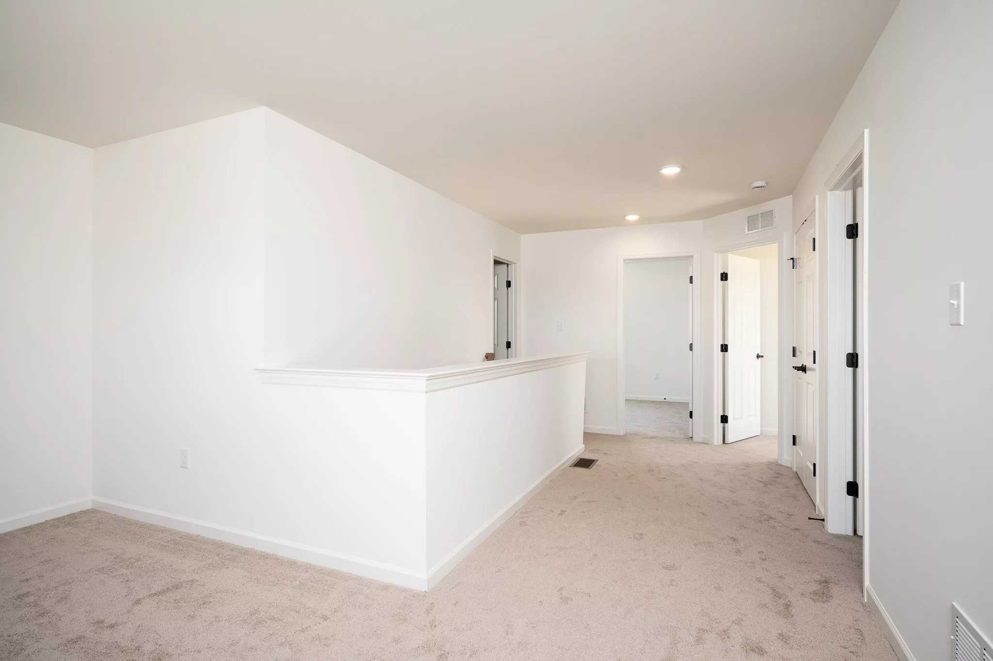 A bright hallway with beige carpet, white walls, recessed ceiling lights, and multiple doors leading to other rooms in a modern home.