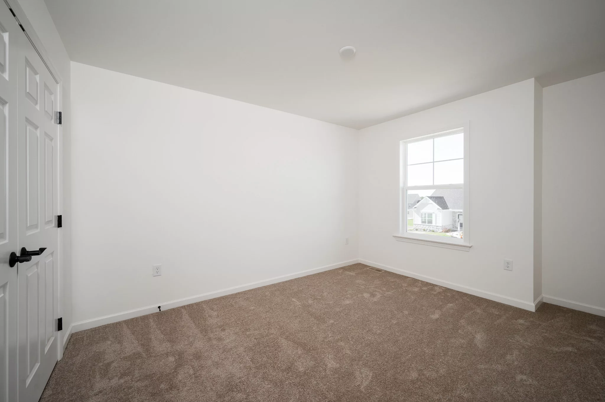 A bright, empty room with white walls, beige carpet flooring, a large window letting in natural light, and a closed white door with black handles.