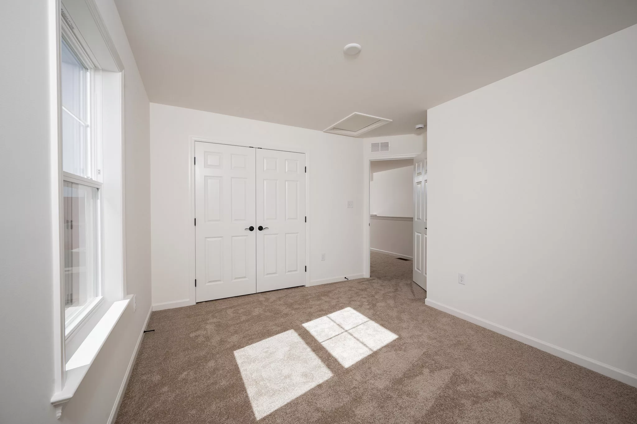 A bright, empty room with beige carpet, white walls, a large window letting in sunlight, and double white closet doors. The ceiling and trim are also white, giving the room a clean, modern look.