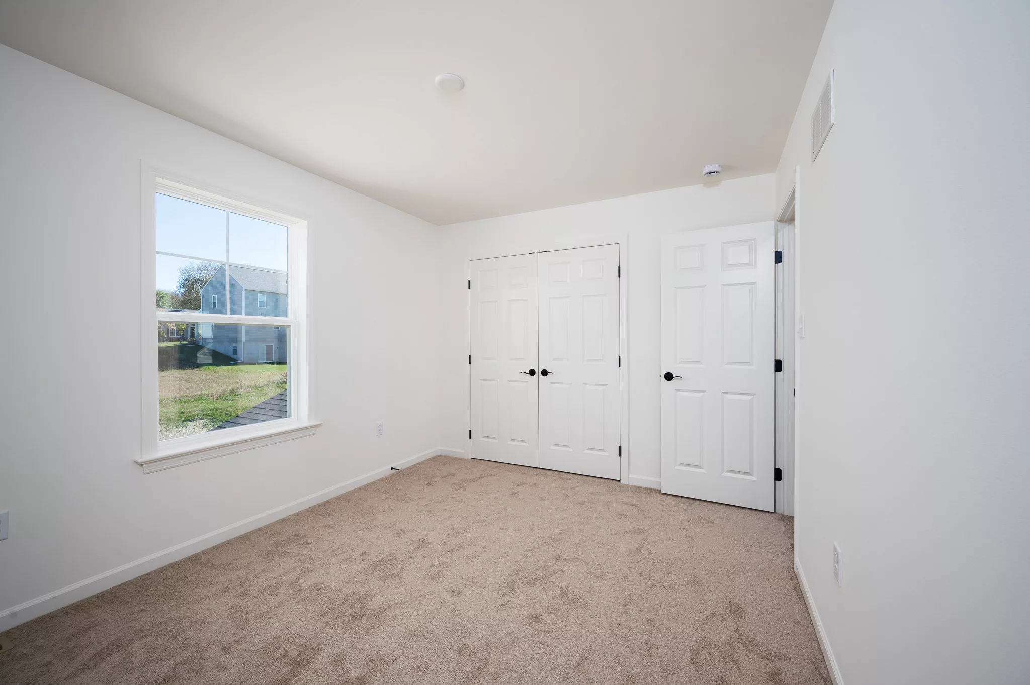 A bright, empty room with beige carpet, white walls, a large window, and two white doors, one being a double closet door. Sunlight shines in through the window, revealing a grassy yard and neighboring house outside.