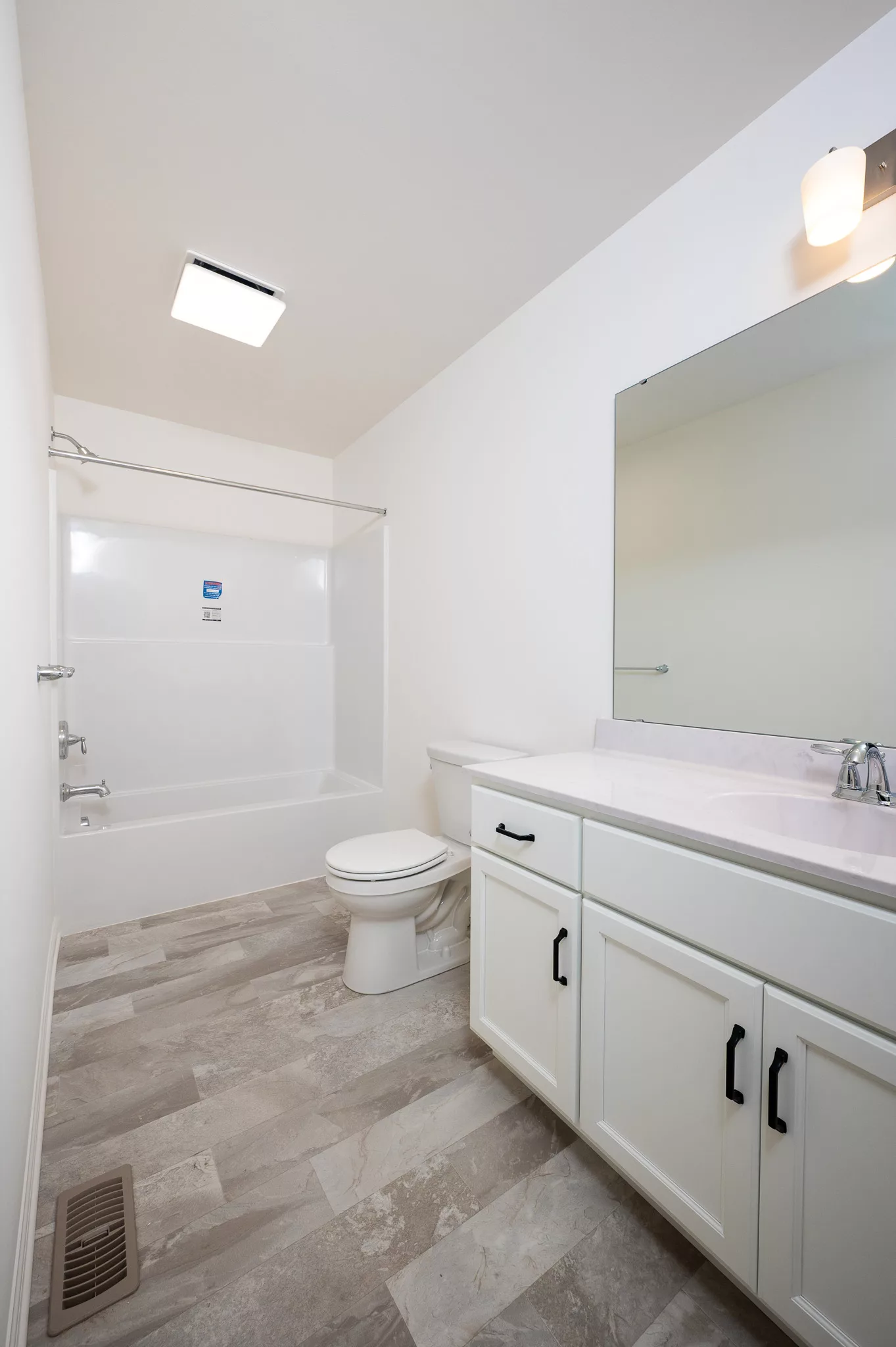 A clean, modern bathroom with white walls, a bathtub-shower combo, a toilet, and a white vanity with a large mirror and under-cabinet storage. The floor is light-colored tile, and a ceiling light is visible.