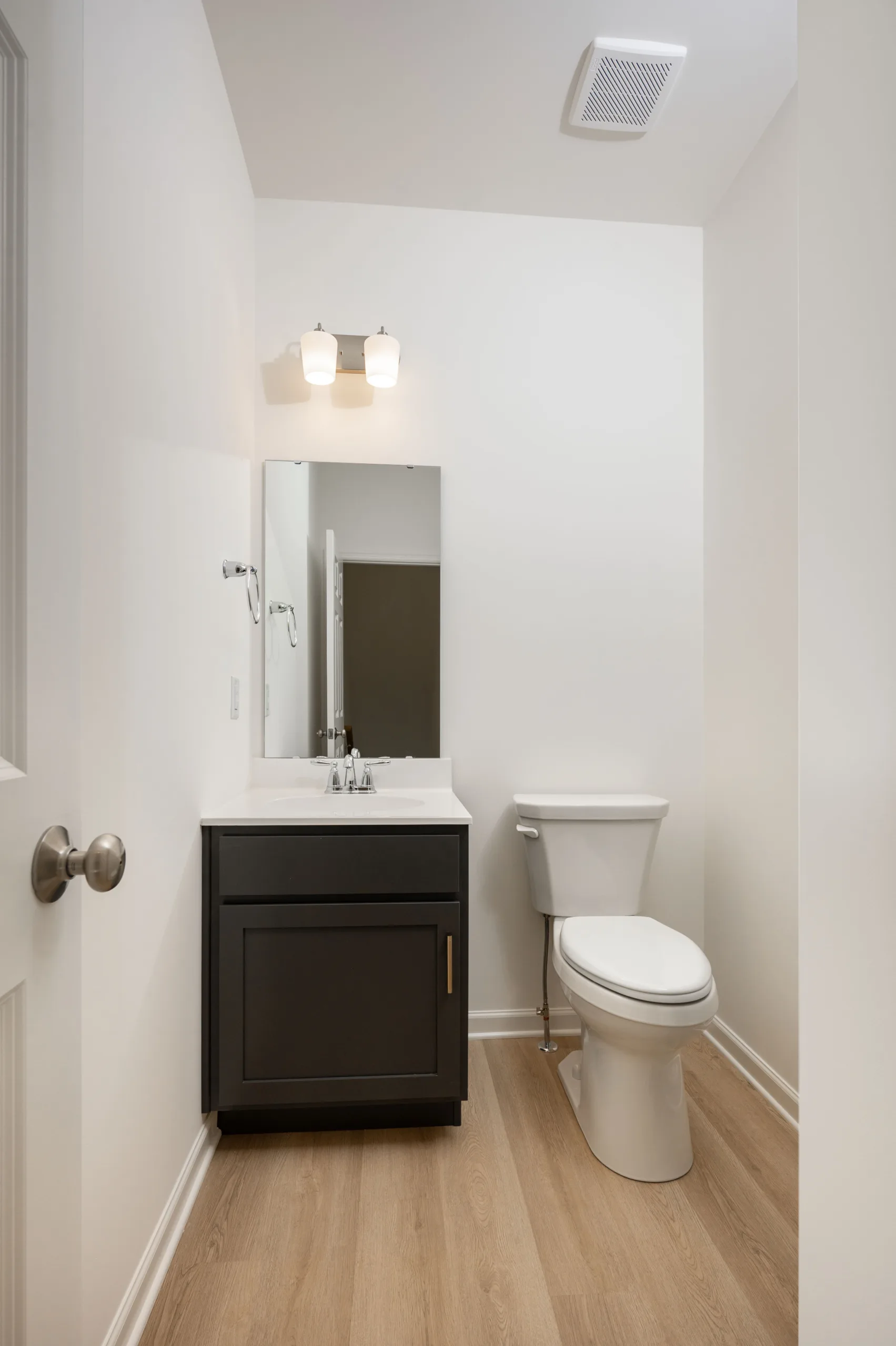 A modern bathroom with a compact sink, mirror, toilet, and light fixture, featuring light wood flooring and white walls.