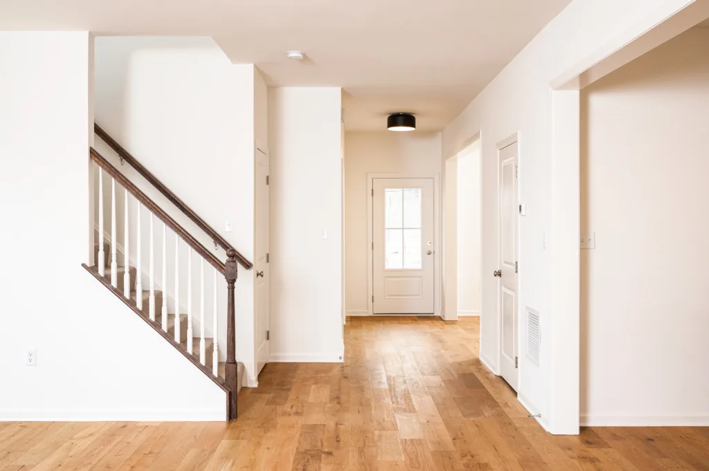 A bright, empty hallway in a modern house with light wood floors, white walls, a staircase with dark railing on the left, and a closed white door with glass panels at the end.