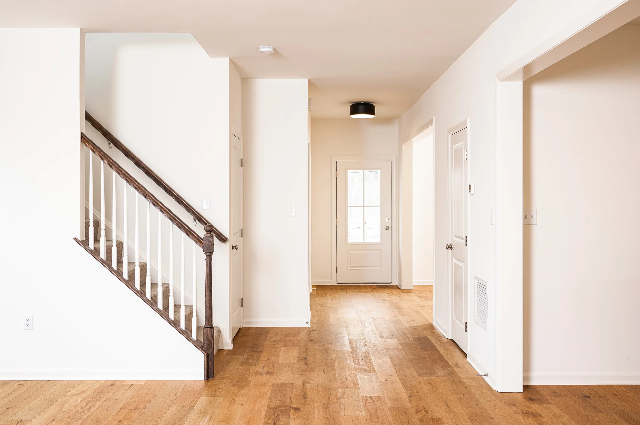 A bright, empty hallway in a modern house with light wood floors, white walls, a staircase with dark railing on the left, and a closed white door with glass panels at the end.