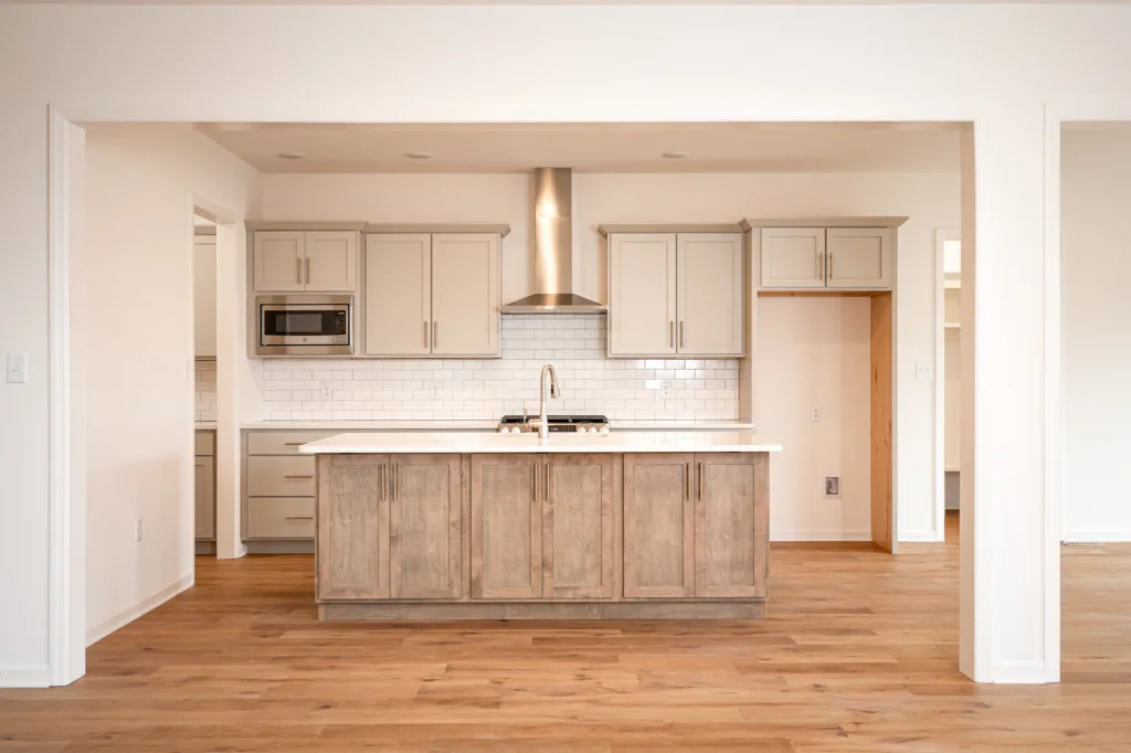 Modern kitchen with light wood flooring, a central island with cabinetry, stainless steel appliances, white tile backsplash, and beige cabinets. The space is bright with clean, neutral colors and minimalist design.