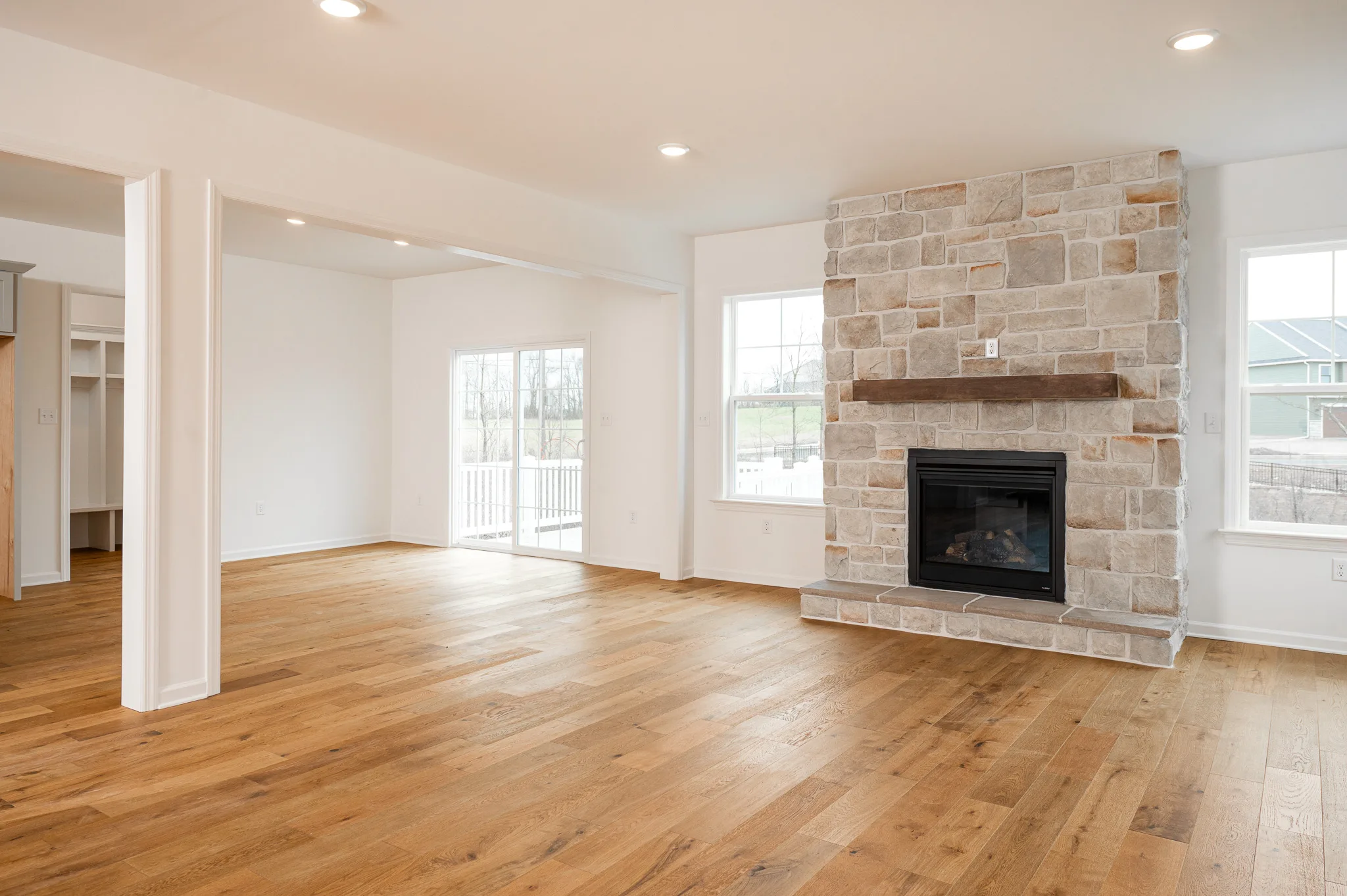 Bright, empty living room with light wood floors, large windows, and a stone fireplace with a wood mantel. Adjacent open area leads to a deck through glass doors. White walls and recessed lighting throughout.