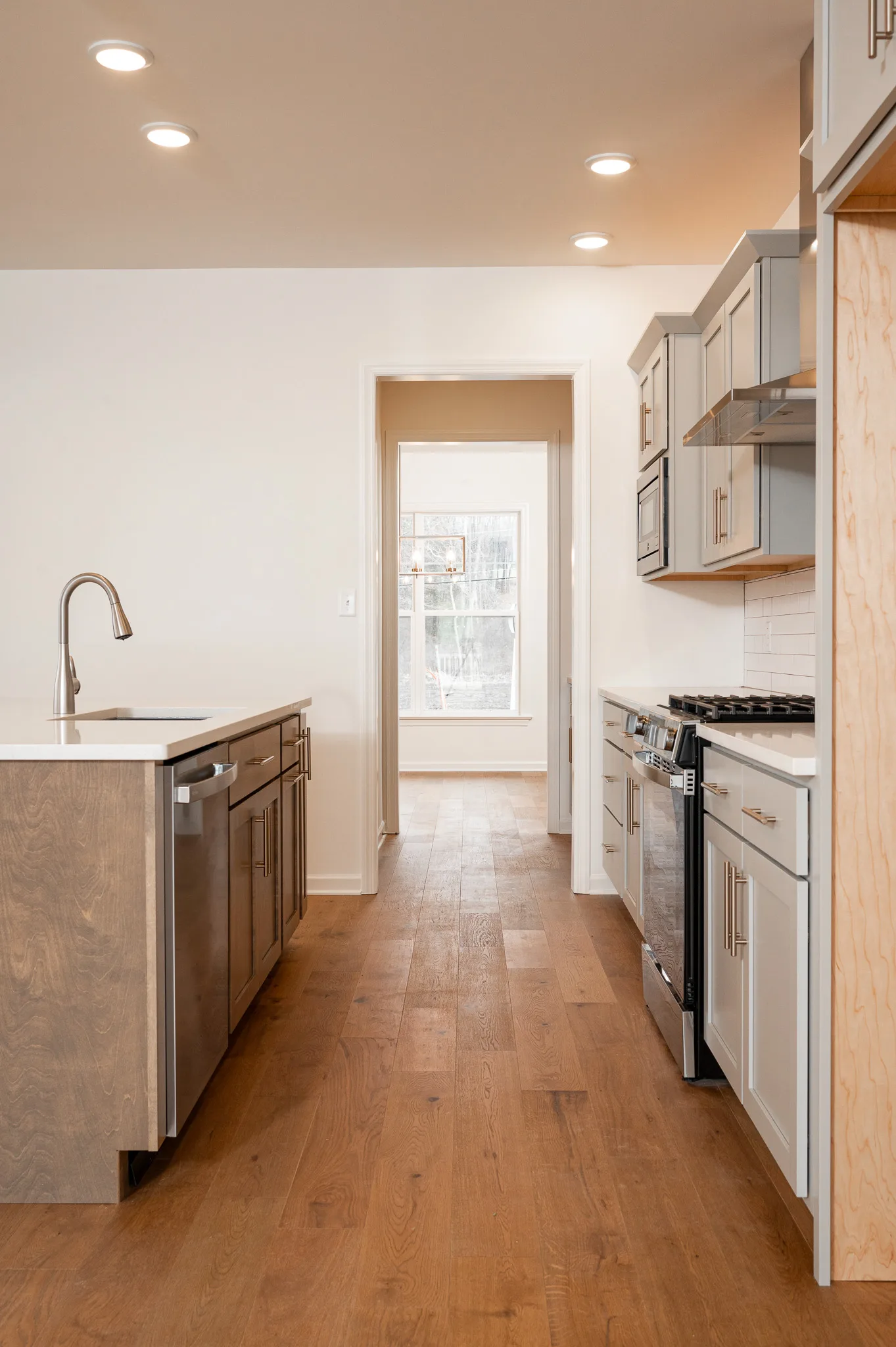 Modern kitchen with light wood floors, white cabinets, a built-in oven, a gas stove, a dishwasher, and an island with a sink, all under recessed ceiling lights. A window is visible at the end of the hallway.