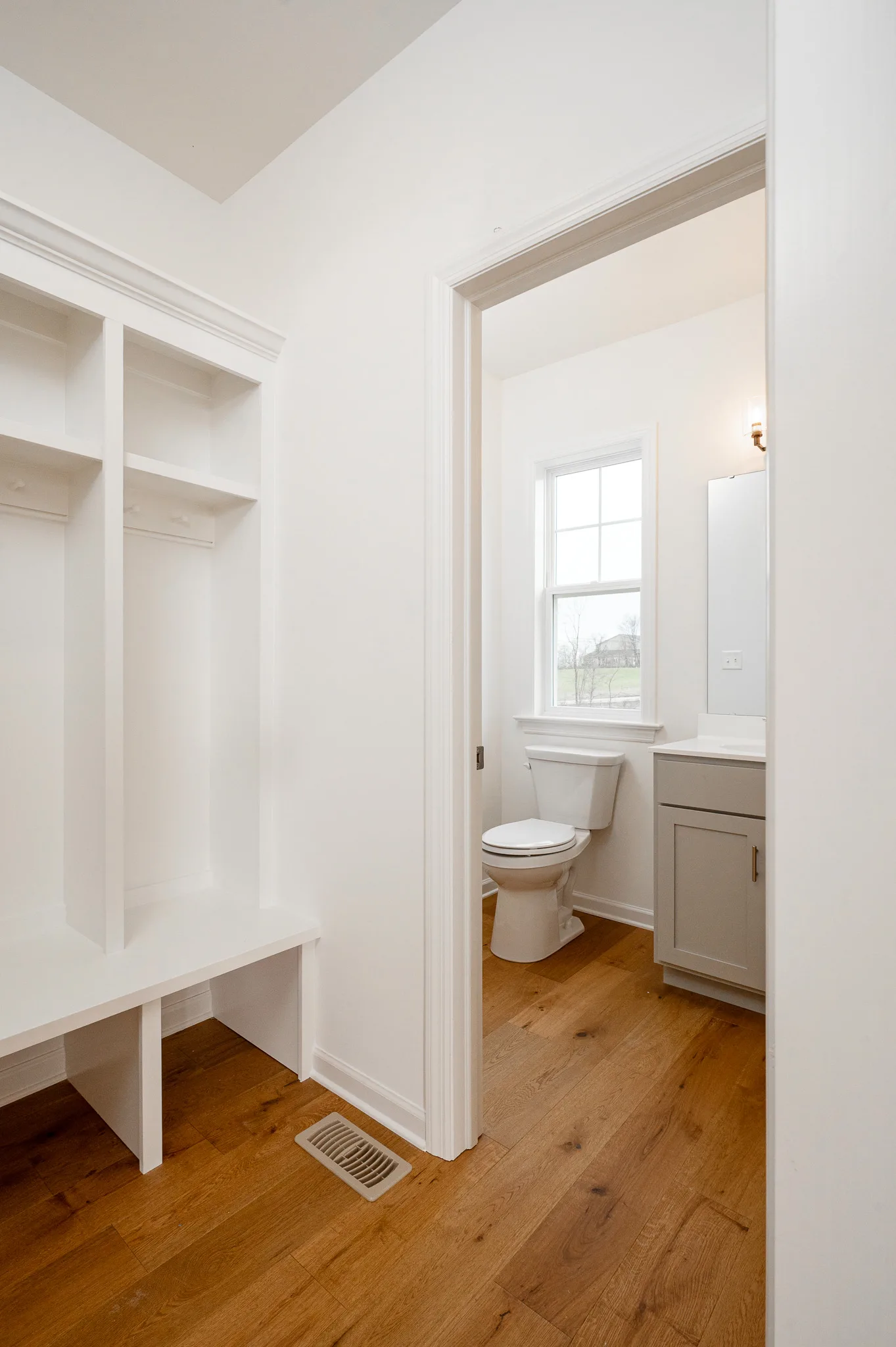 A bright, modern bathroom with white walls, wood flooring, a small window, a toilet, and a light gray vanity. Outside the bathroom is a built-in white storage bench with open shelves.
