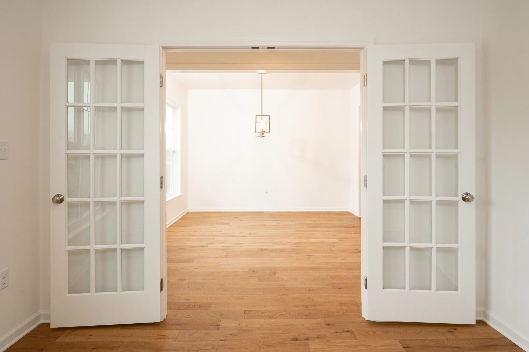 Double glass French doors open into a bright, empty room with light wood floors, white walls, and a modern ceiling light fixture. Natural light comes in through large windows.