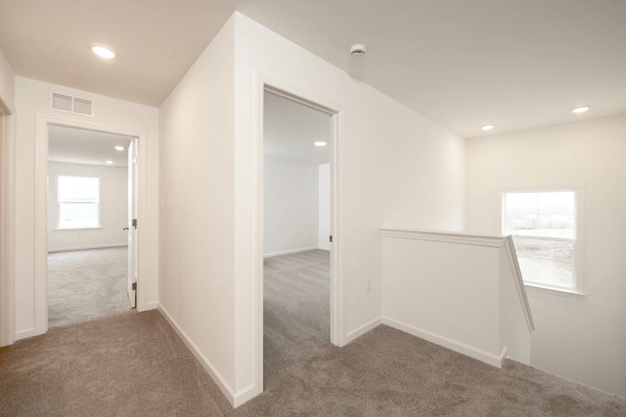 Second-floor hallway with beige carpet, white walls, two open doorways leading to rooms, and a stairway with a window providing natural light.